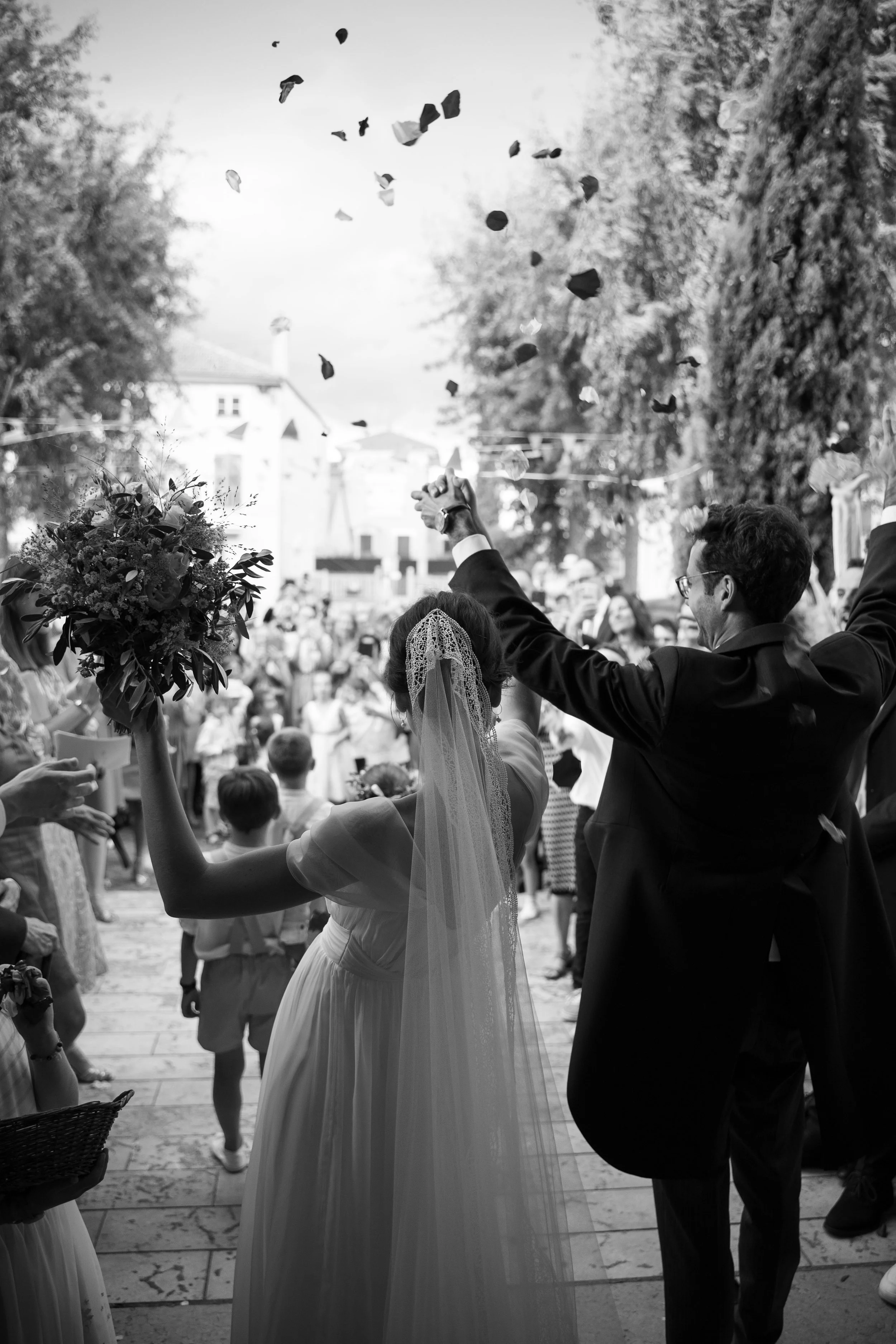 Photo noir et blanc d'un mariage avec des invités, une mariée tenant un bouquet et un couple dansant. Des enfants et d'autres invités en arrière-plan, avec des arbres et des bâtiments visibles.