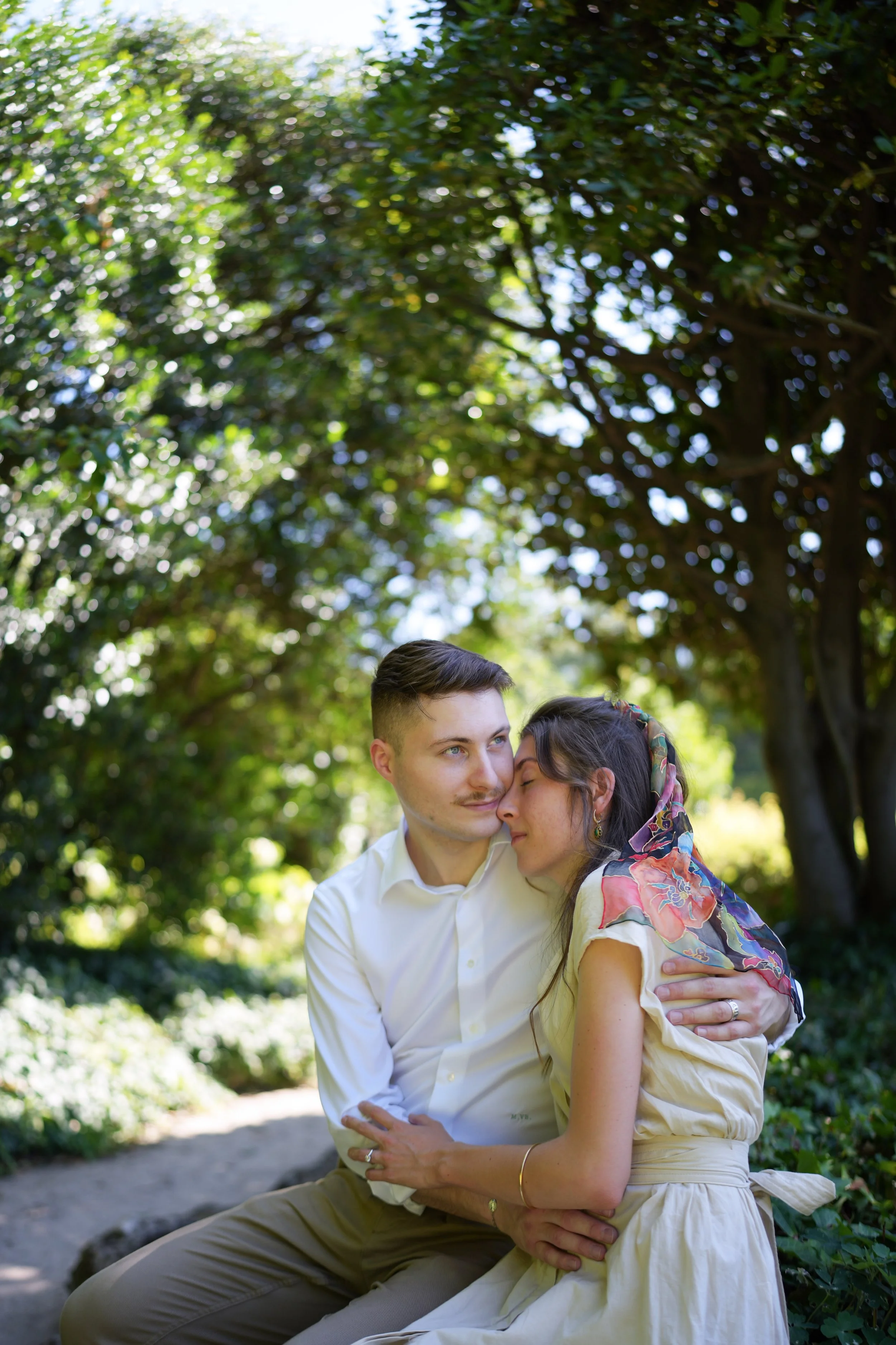 Un couple s'embrasse tendrement dans un parc, sous un arbre avec des feuilles vertes, lors d'une journée ensoleillée.
