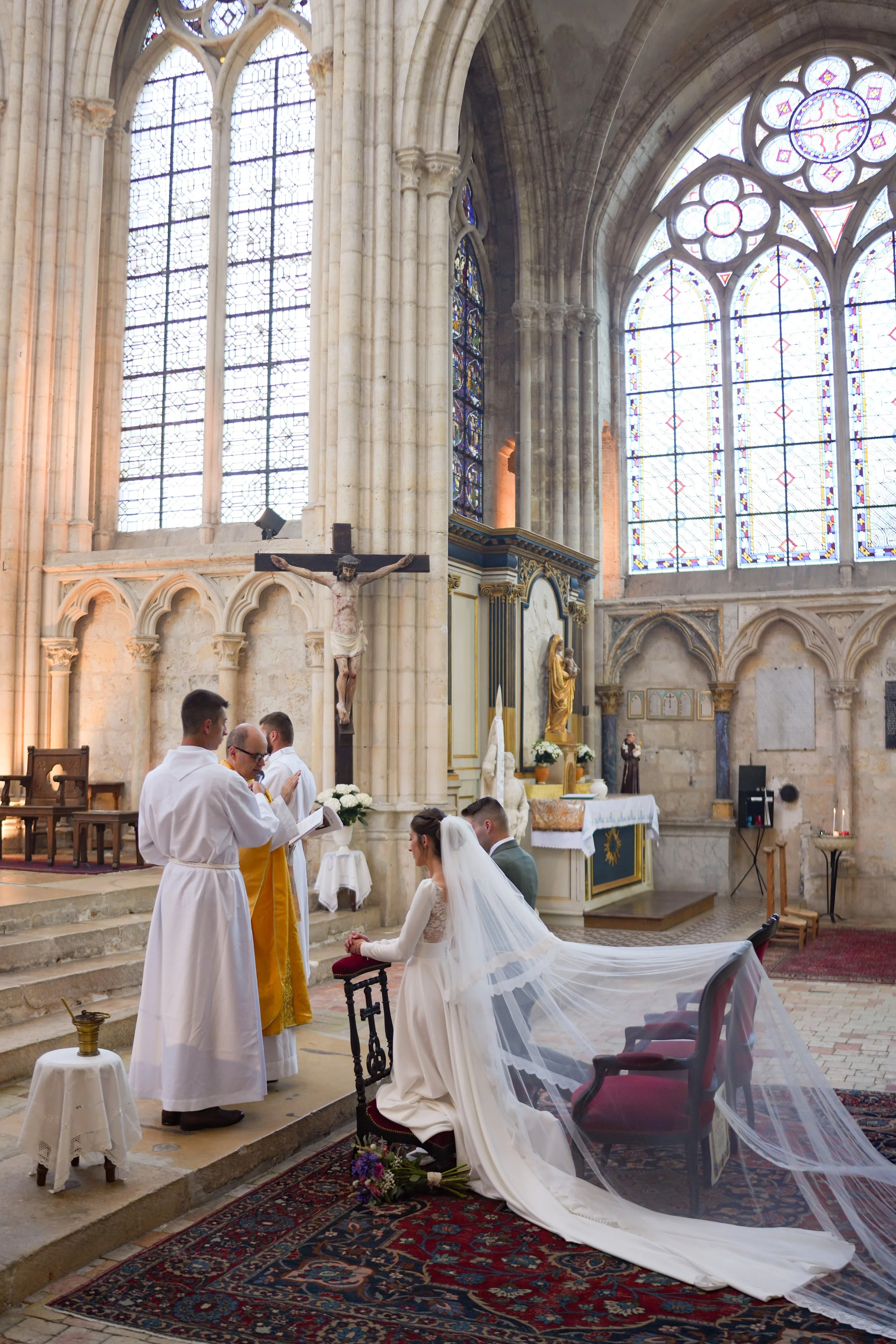 Un mariage religieux dans une grande cathédrale, avec des personnes en prière et un couple de mariés à genoux devant un prêtre, un crucifix et des vitraux colorés en arrière-plan.