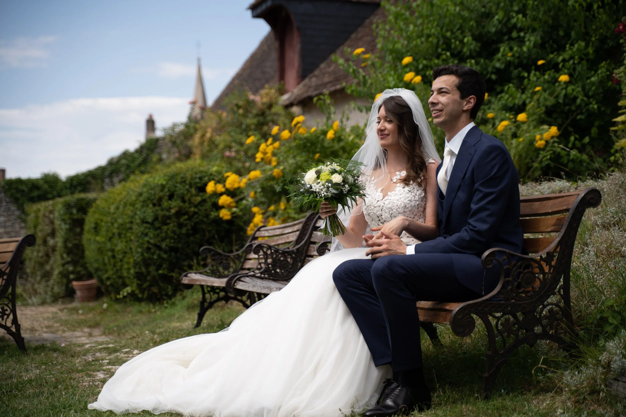Un couple de mariés assis sur un banc en plein air, entouré de verdure et de fleurs jaunes. Elle porte une robe de mariage blanche avec une voile, il porte un costume bleu foncé. La femme tient un bouquet de fleurs. Le ciel est partiellement nuageux.
