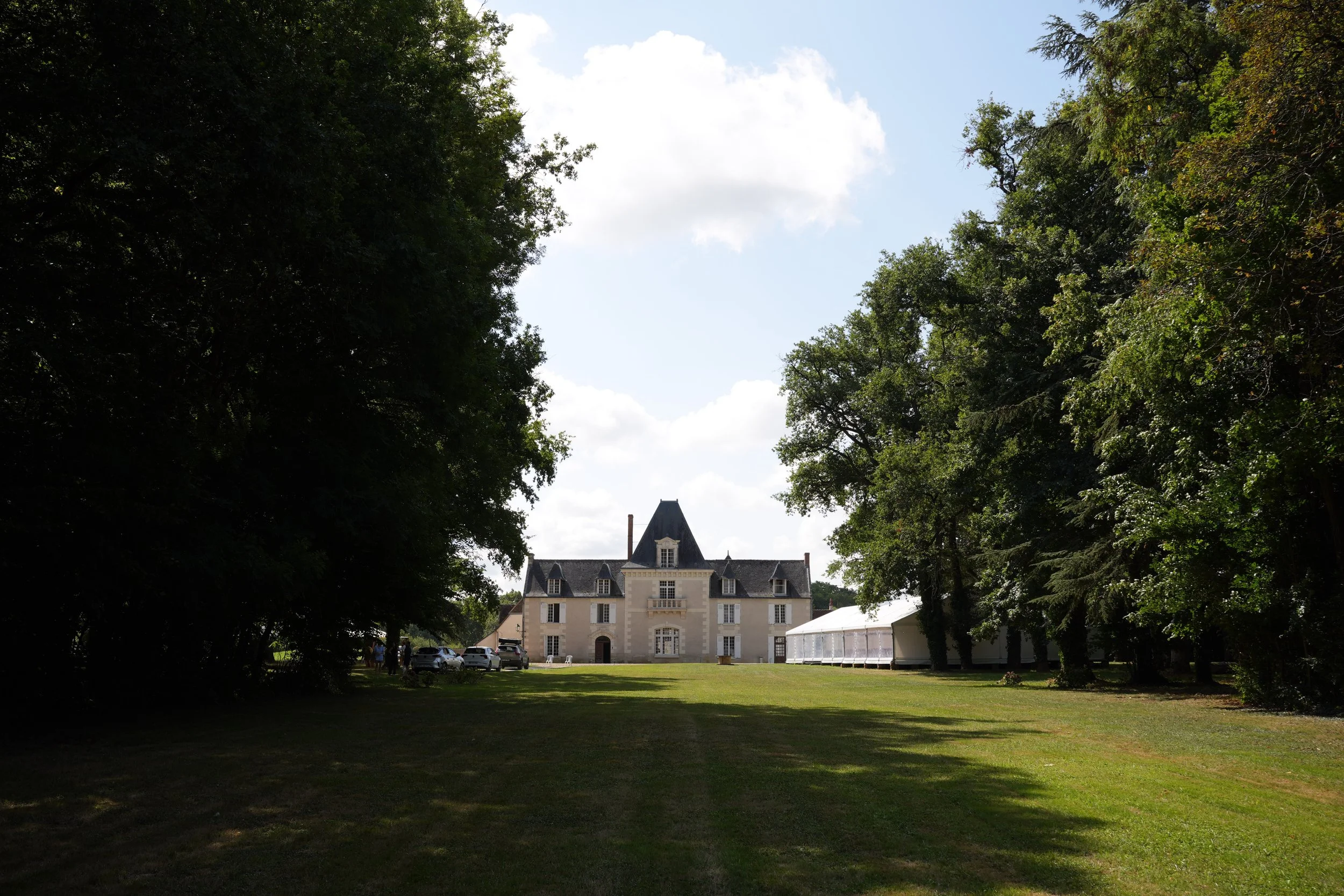 Un grand bâtiment de style château avec plusieurs toits pointus, entouré d'arbres verts, dans un parc en extérieur avec des voitures garées devant