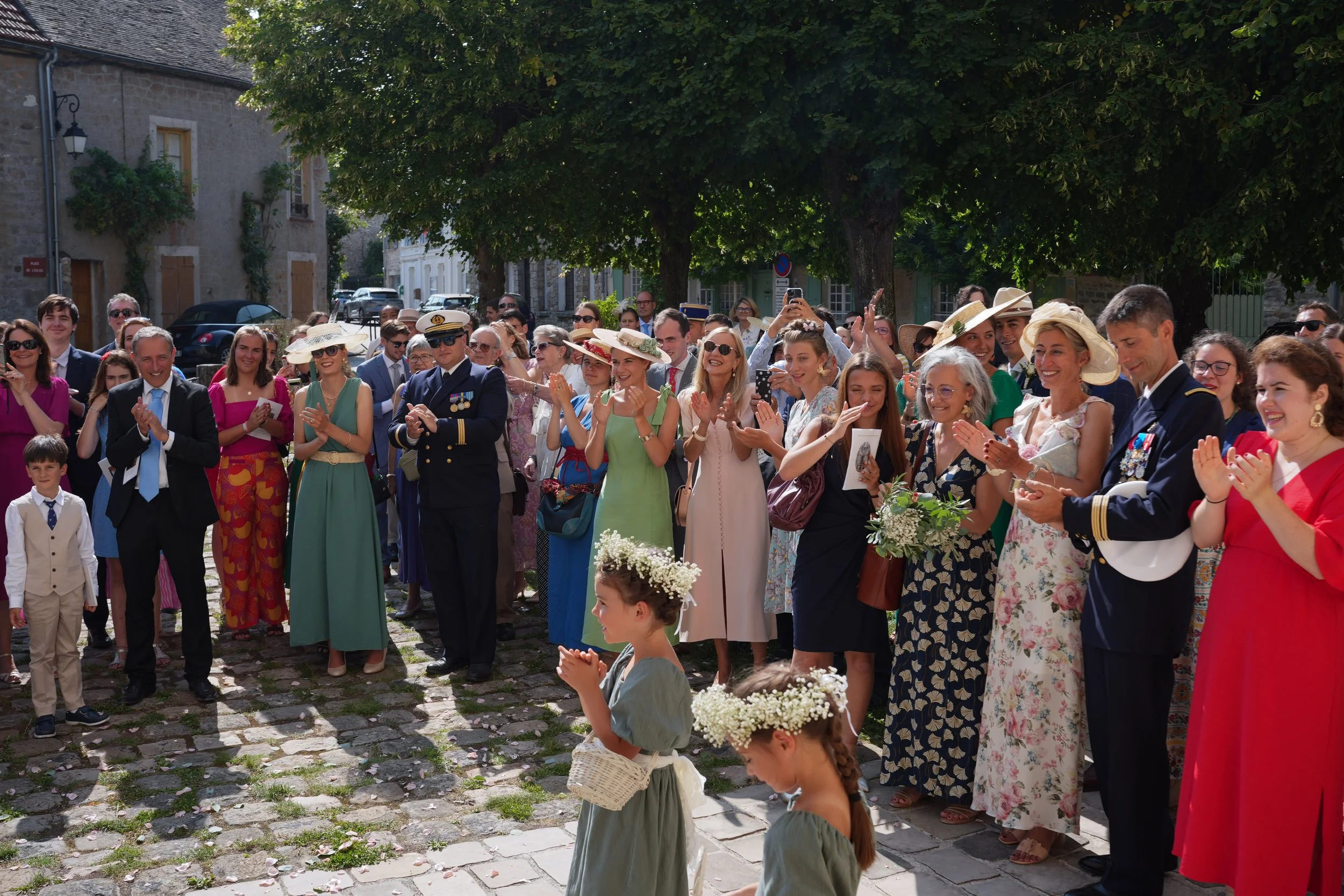 Un groupe de personnes lors d'une célébration en plein air, portant des vêtements élégants et des chapeaux, applaudissant sous un arbre feuillu dans un village.