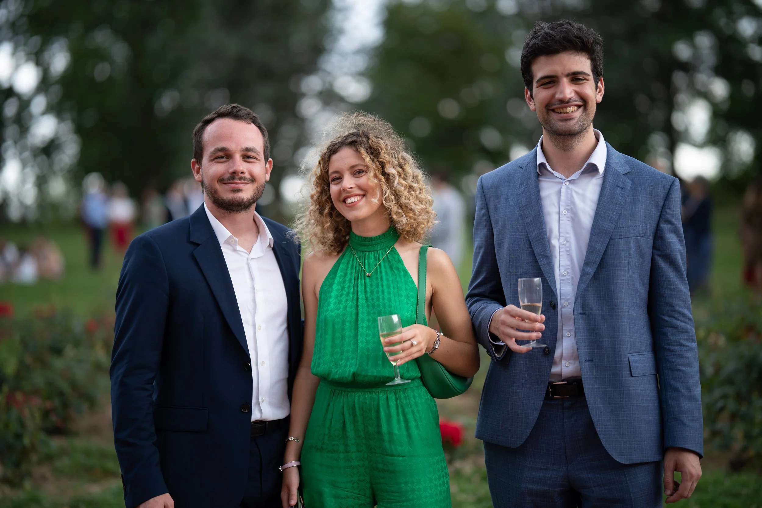 Trois personnes habillées formellement, deux hommes et une femme, posent ensemble en extérieur lors d'une célébration ou événement social, tenant des flûtes à champagne, avec un arrière-plan de nature et autres invités.