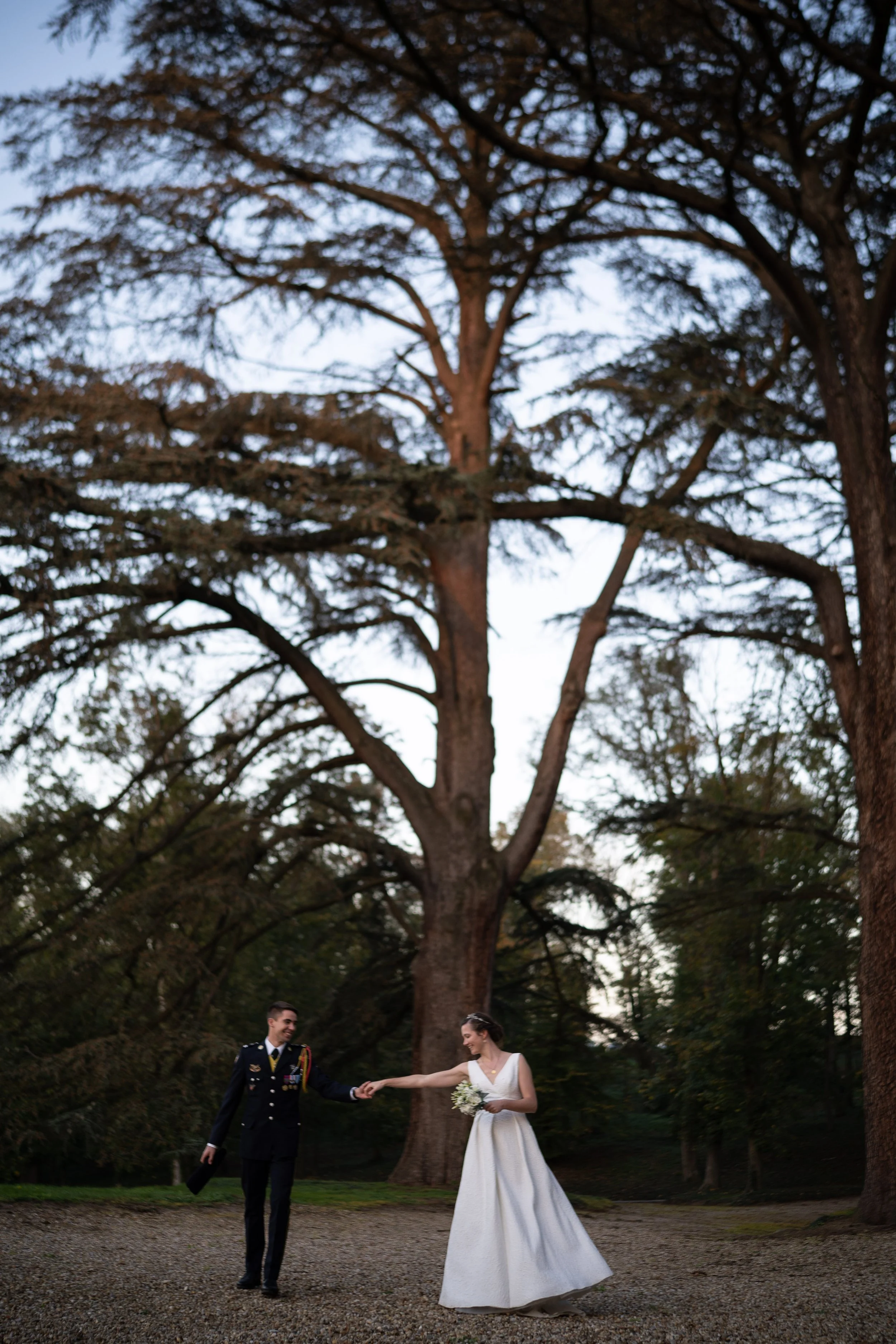 Un couple habillé pour un mariage, la femme en robe blanche et le homme en uniforme militaire, se tenant par la main sous un grand arbre lors de la soirée.
