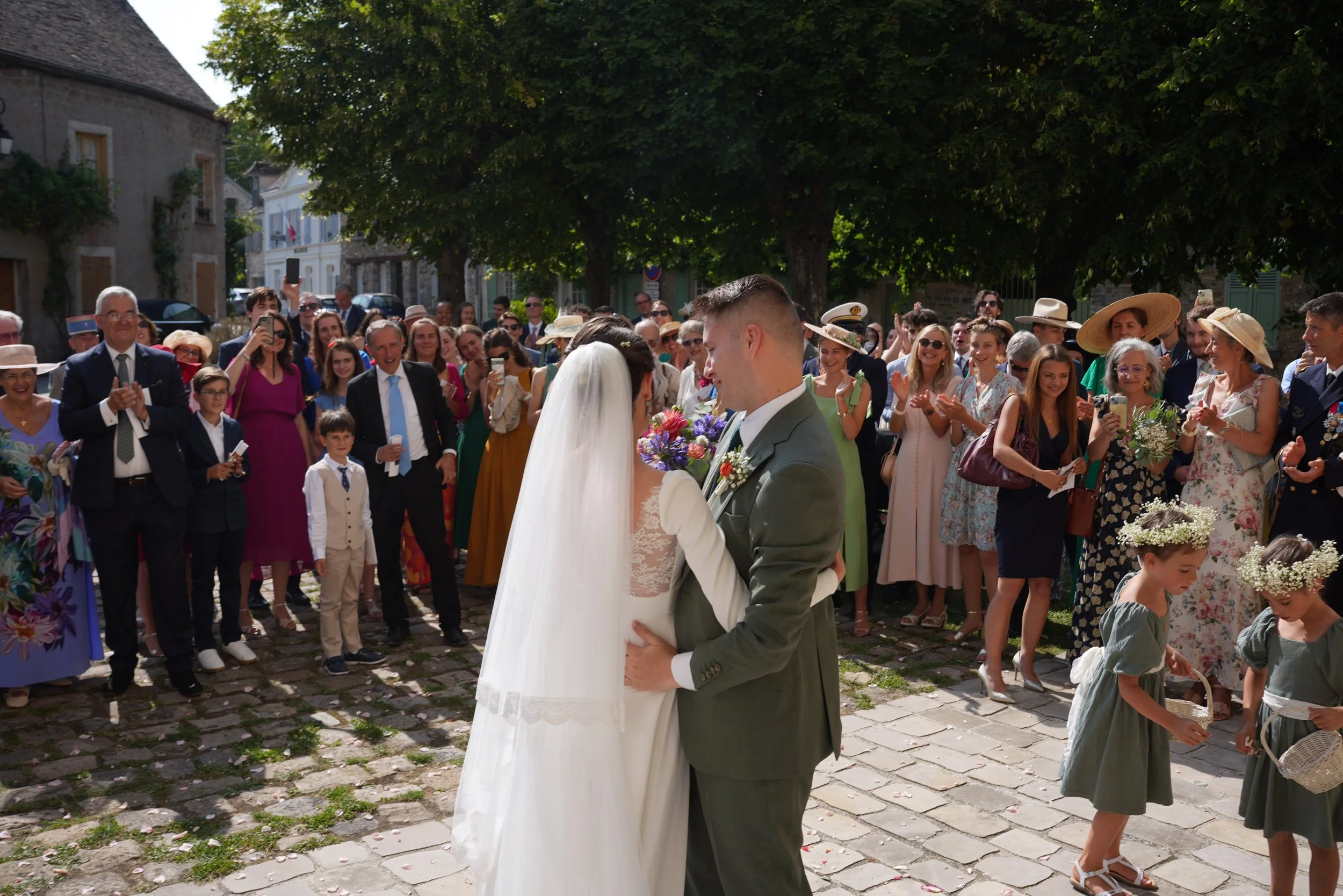 Un couple de mariés s'embrassant lors de leur mariage, entouré d'une foule d'invités riant et prenant des photos, en plein air sous de grands arbres.