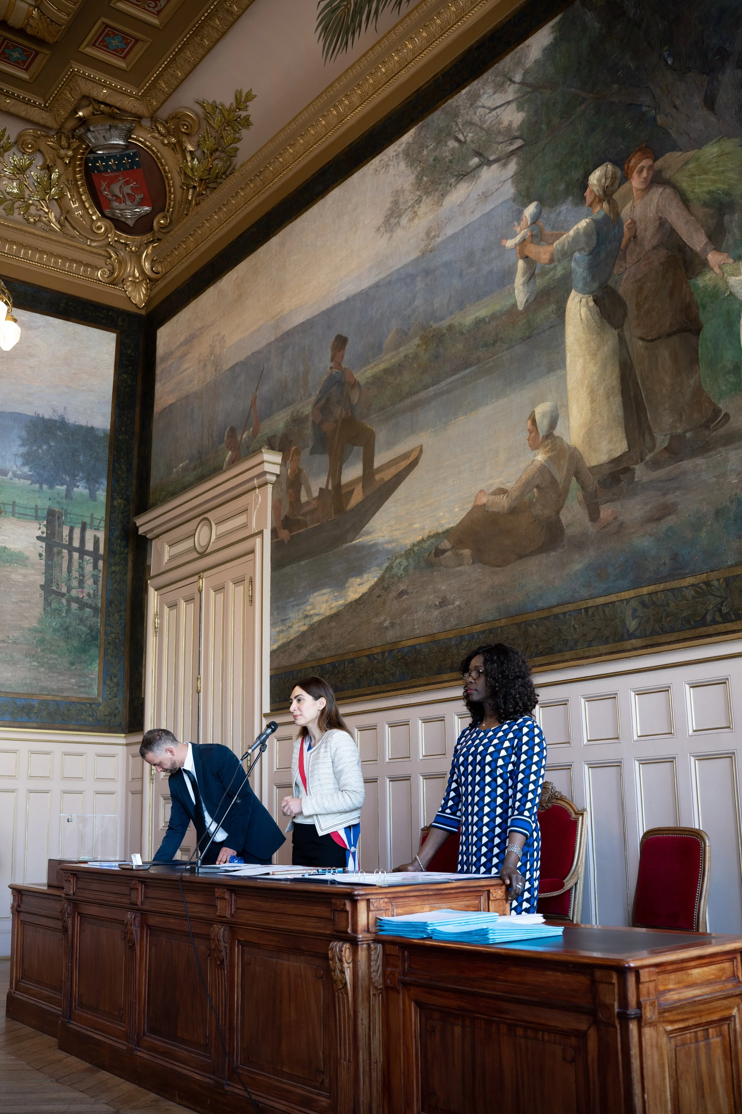 Une scène de réunion dans une salle ornée de tableaux et de décorations anciennes, avec des personnes debout derrière une grande table en bois, des documents et un microphone, et un grand tableau mural représentant des femmes en train de pêcher au bo