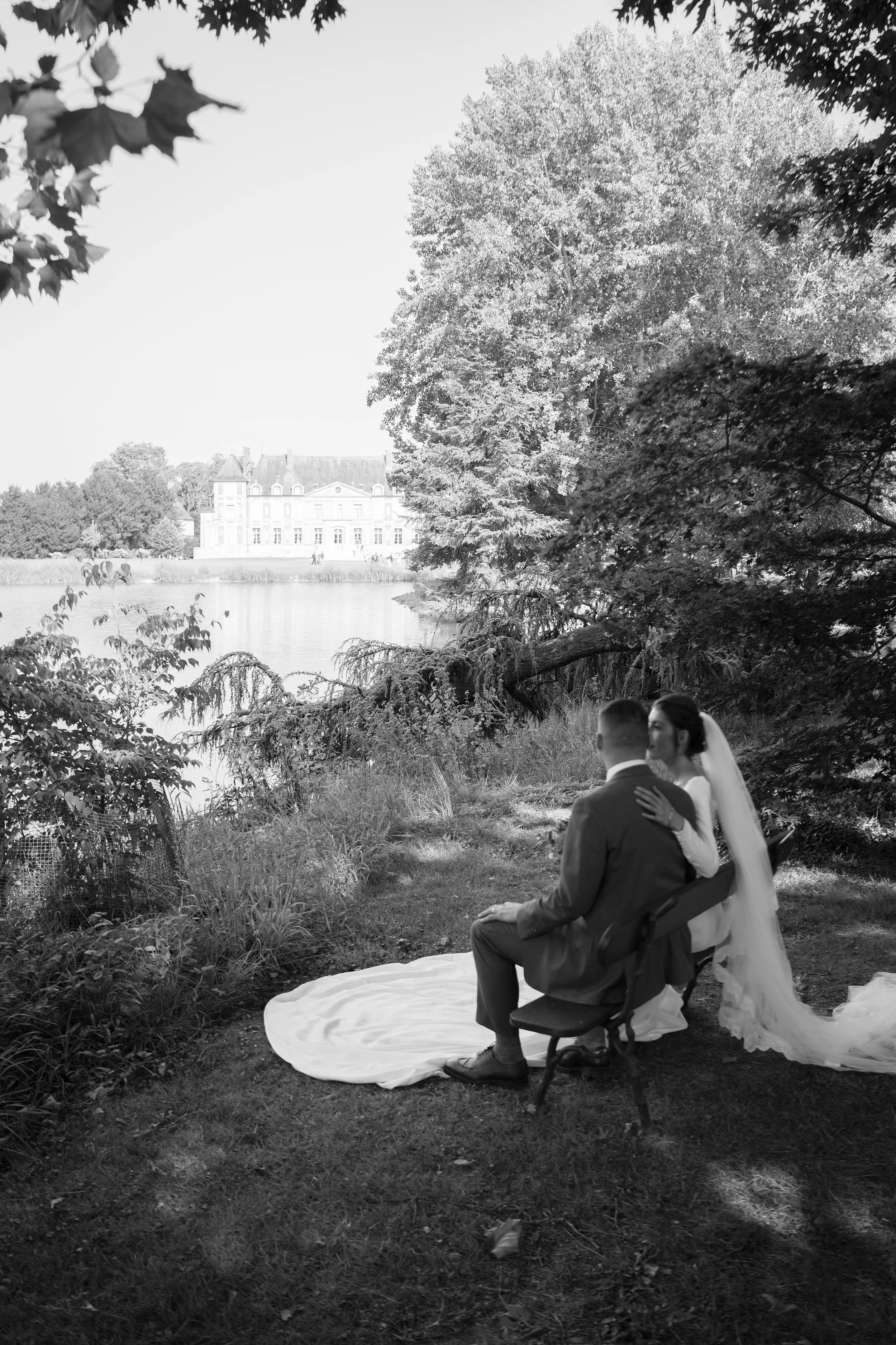 Un couple lors d'un mariage, assis dans la nature près d'un lac, avec un château en arrière-plan, en noir et blanc.