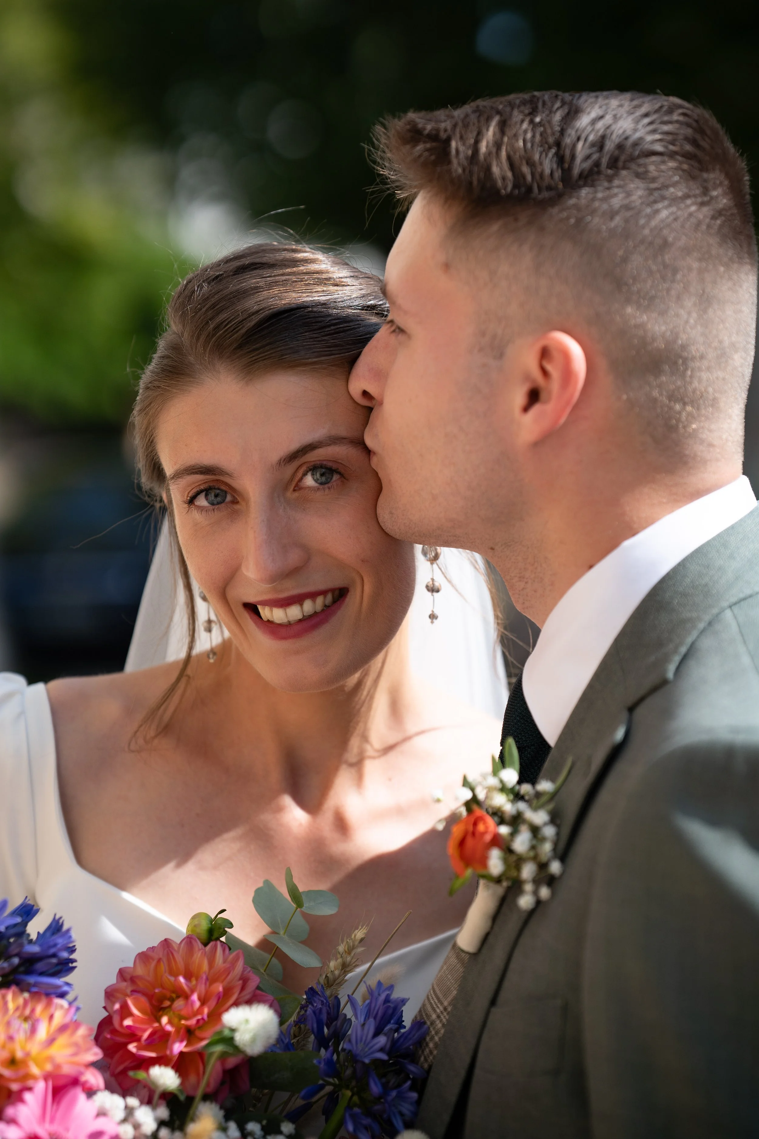 Un couple de mariage, la femme avec un sourire, le regard tourné vers l'objectif, et le mari en train de l'embrasser sur le front. La femme tient un bouquet de fleurs colorées. L'arrière-plan est flou, suggérant une scène extérieure.