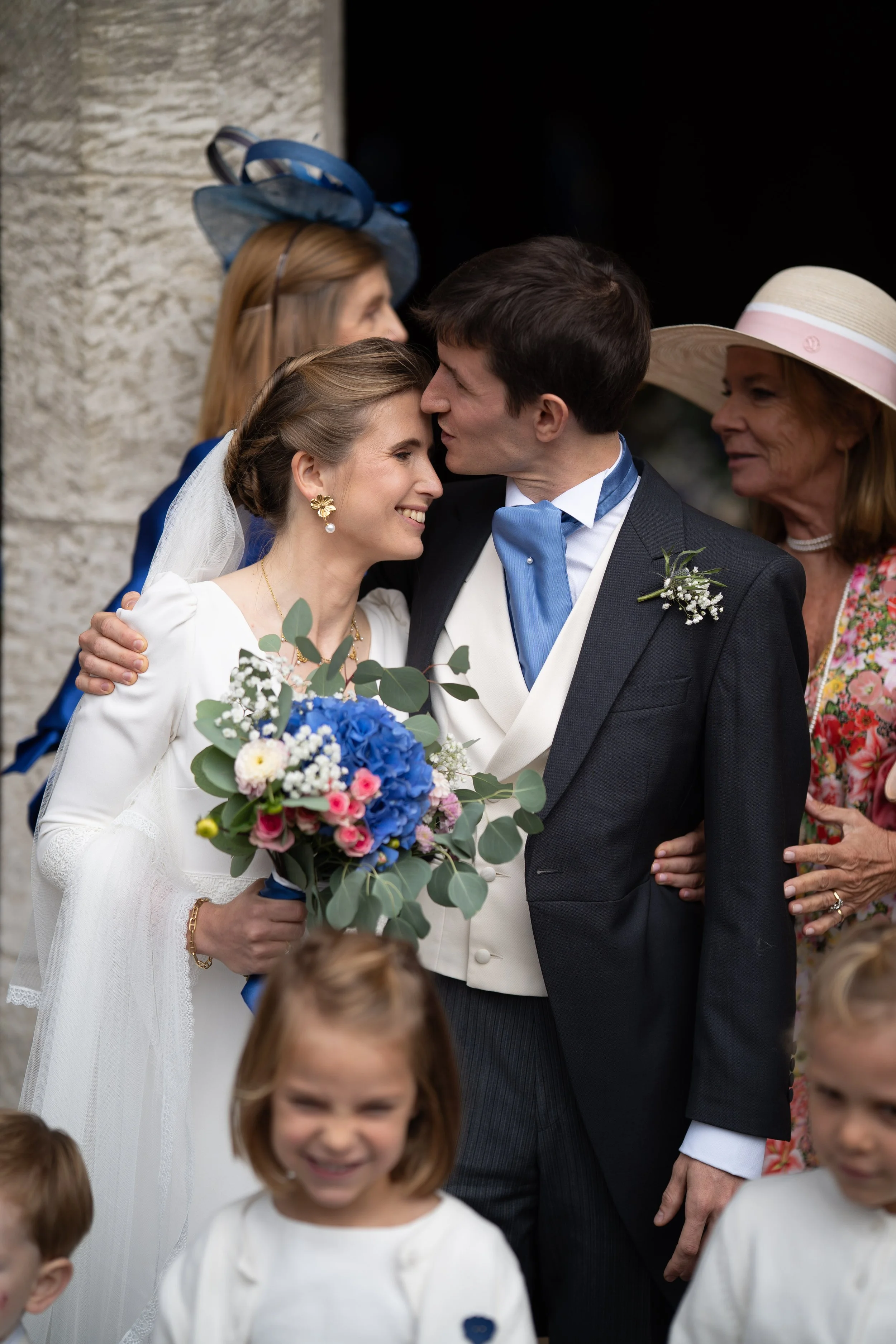 Un couple de mariés s'embrasse tendrement lors d'une cérémonie. La mariée porte une robe blanche, un voile et des boucles d'oreilles en or avec un pendentif en perle, et tient un bouquet de fleurs colorées. Le marié porte un costume noir avec un nœud