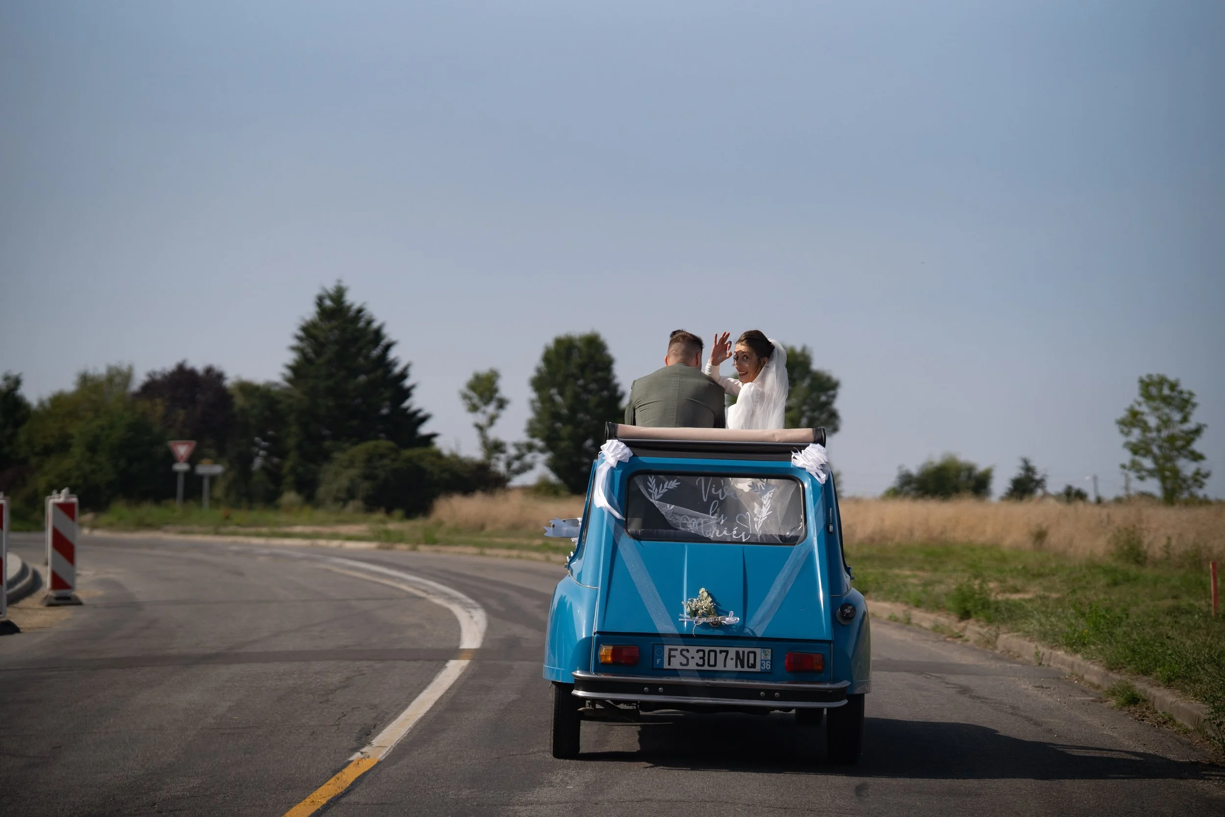 Un couple marié à bord d'une petite voiture bleu ciel roulant sur une route de campagne lors d'un mariage.