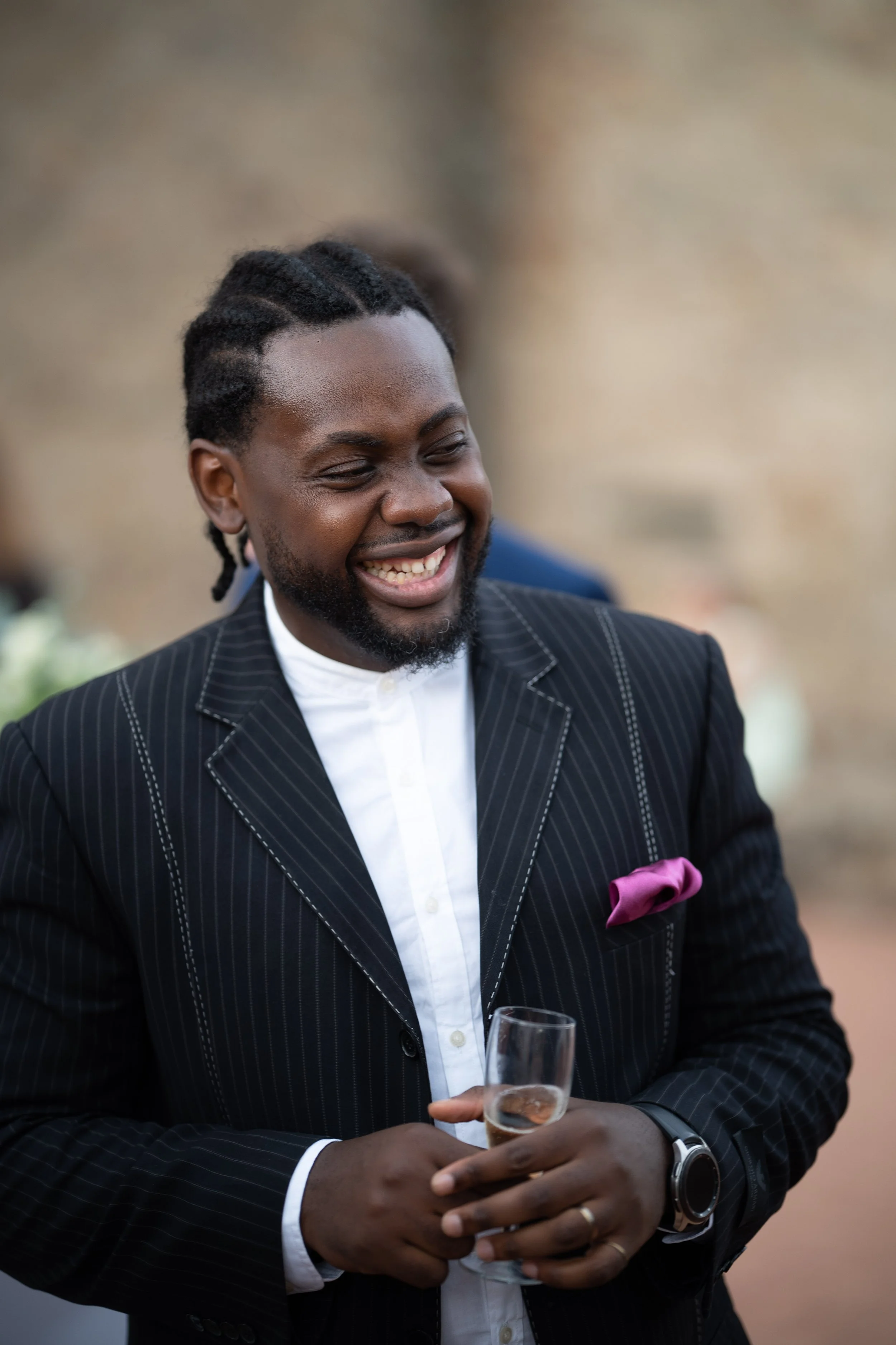 Un homme souriant, en costume noir avec des rayures blanches, tenant un verre de champagne dans la main, lors d'un événement social ou une fête.