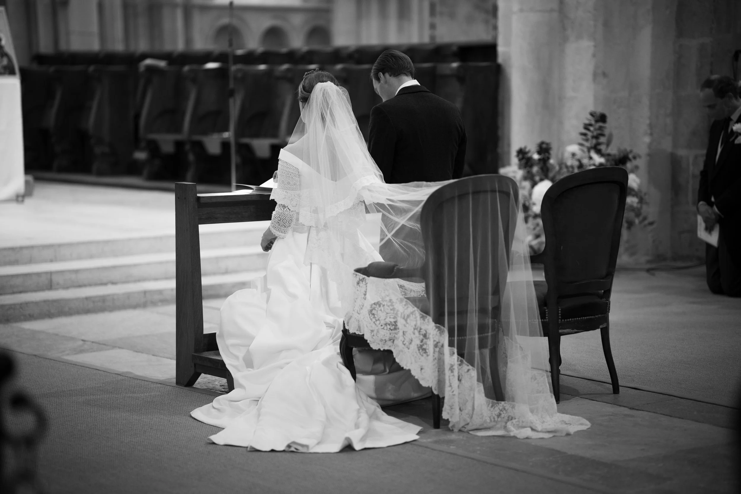 Un couple de mariés prie dans une église, la mariée en robe blanche avec voile et le marié en costume noir, assis devant l'autel.