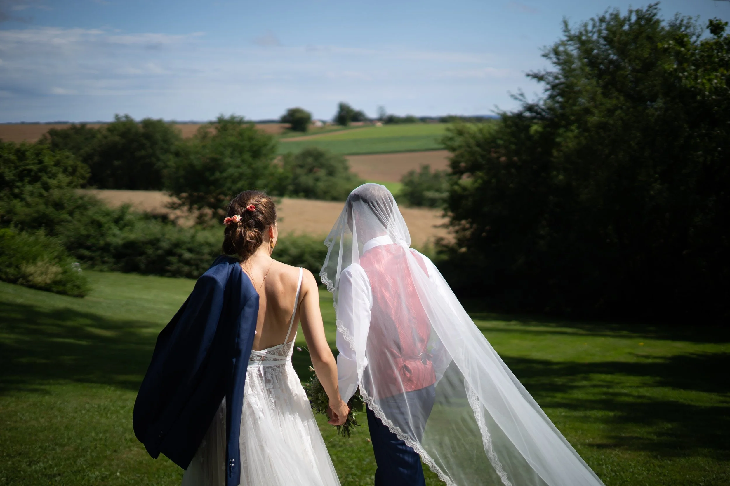 Deux femmes en robes de mariée et avec un voile tiennent la main dans un paysage rural avec champs et arbres sous un ciel bleu.