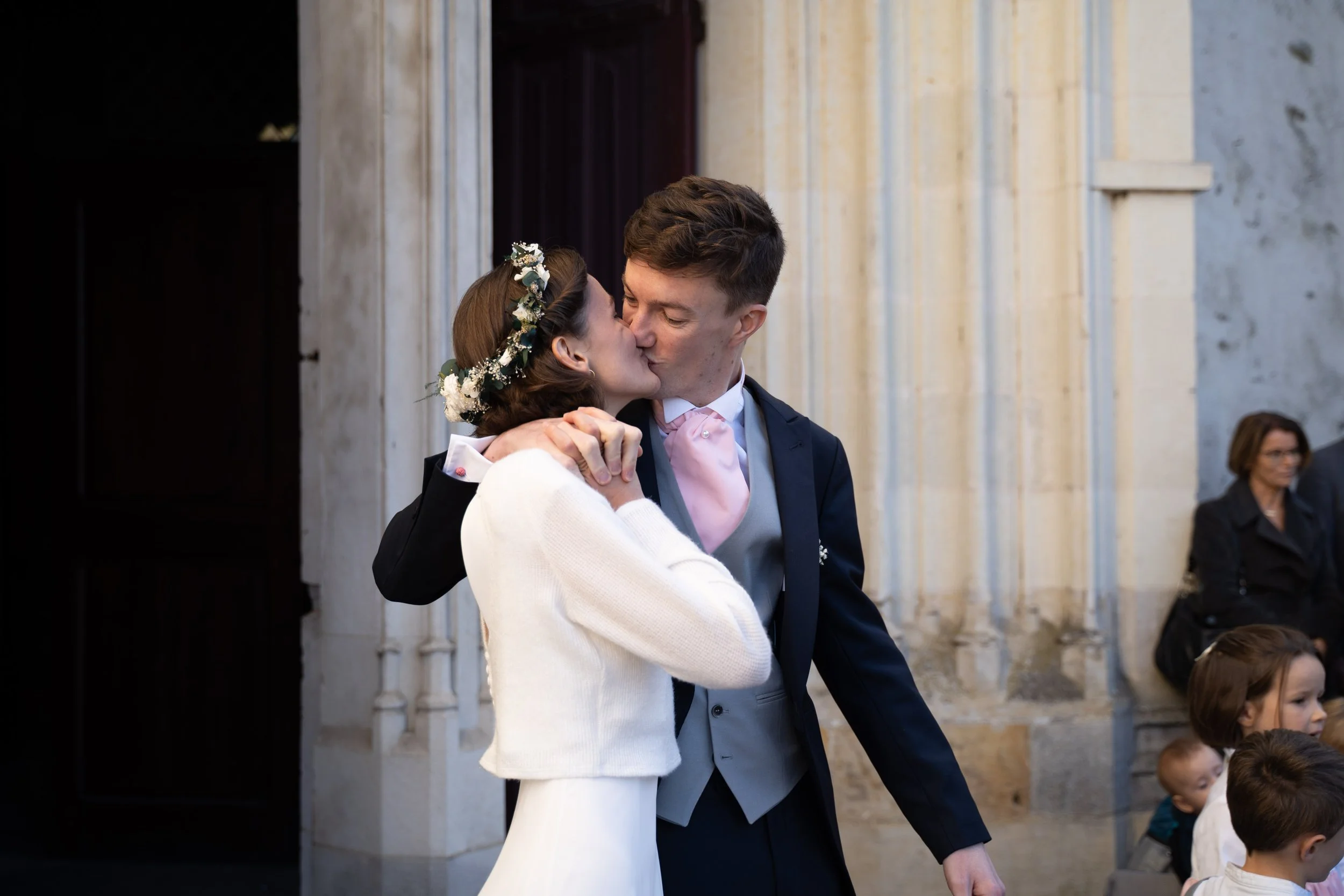 Un couple marié s'embrasse lors de leur cérémonie dans un lieu architectural en pierre, avec des invités en arrière-plan.