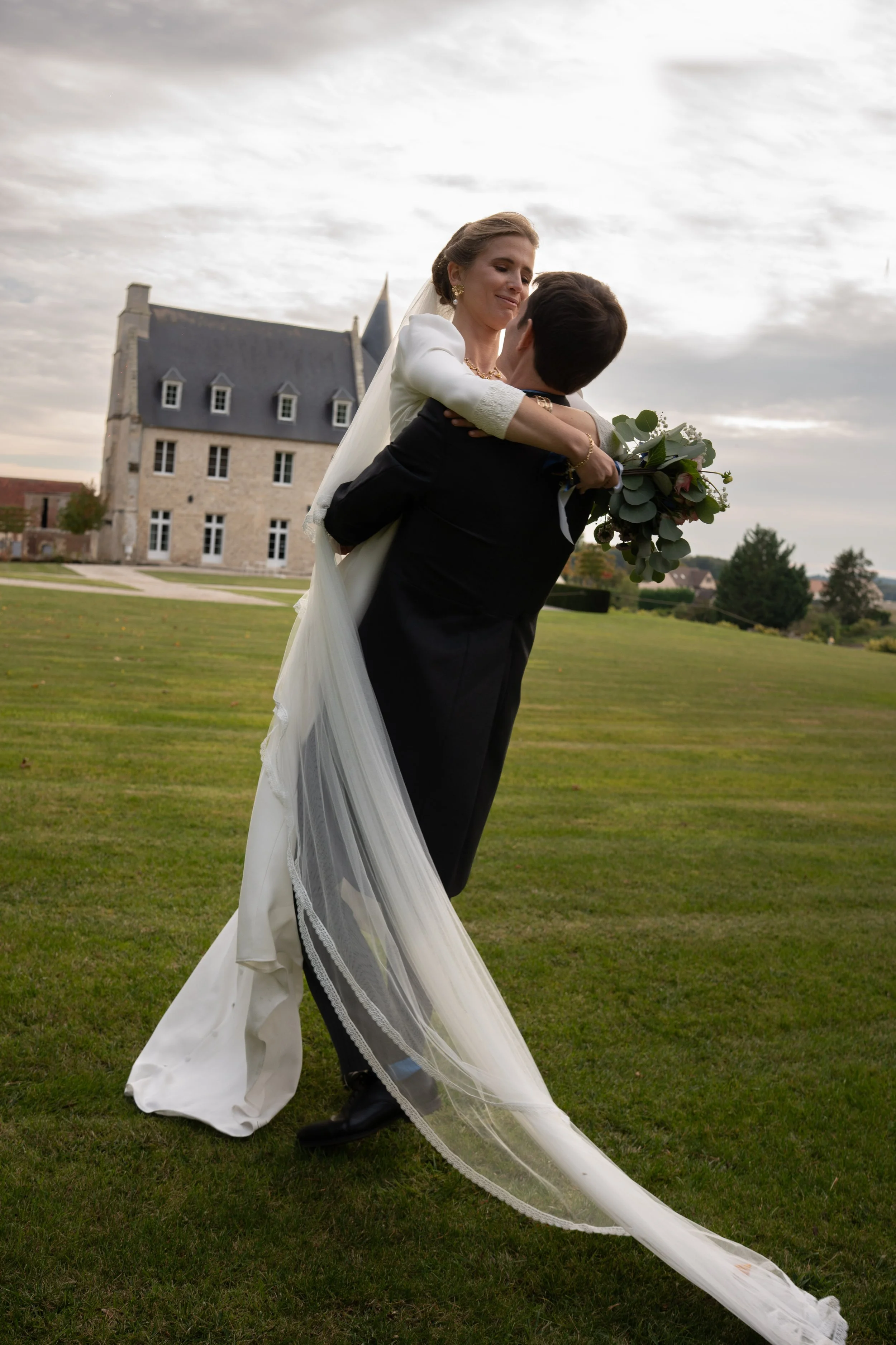 Un couple de mariés, la femme en robe blanche avec un voile et le homme en costume noir, s'enlace dans un jardin avec un grand bâtiment en pierre en arrière-plan, sous un ciel nuageux.
