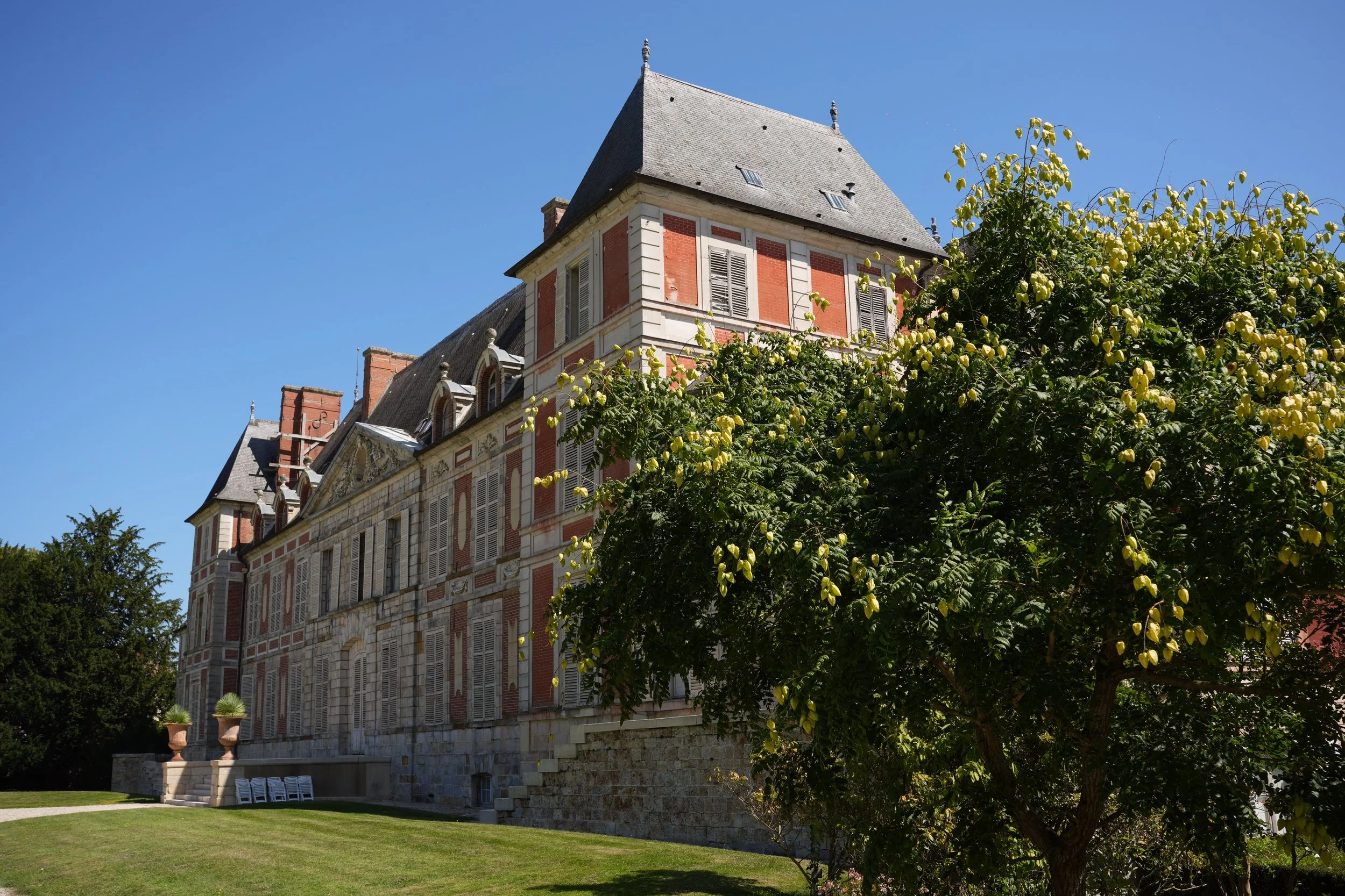 Une grande bâtisse historique en pierre et brique avec des fenêtres à volets et un toit en pente, entourée d'arbres feuillus, sous un ciel bleu clair.