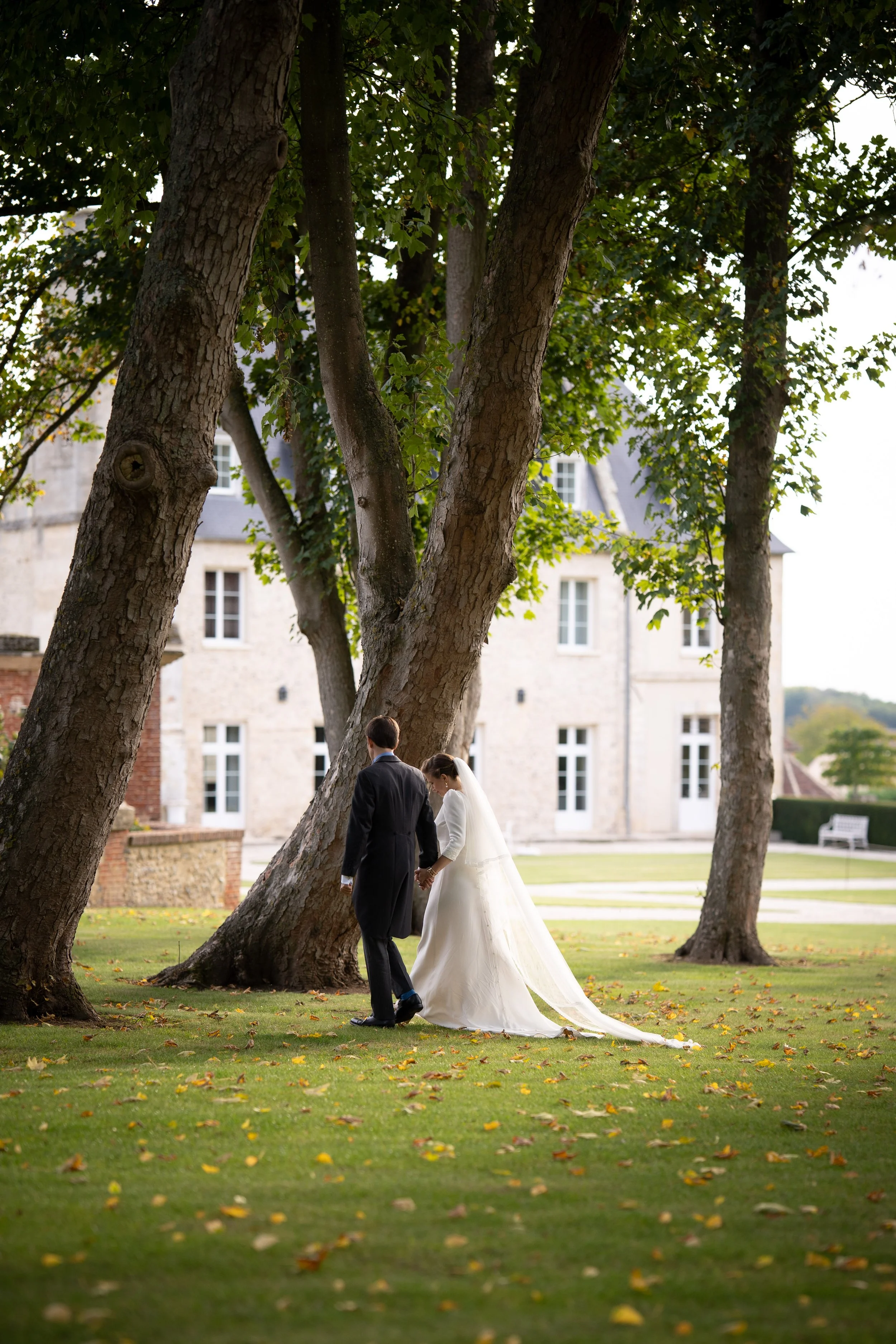 Un couple de mariés marchant dans un parc, entouré d'arbres aux feuilles jaunes, avec un bâtiment en pierre en arrière-plan.
