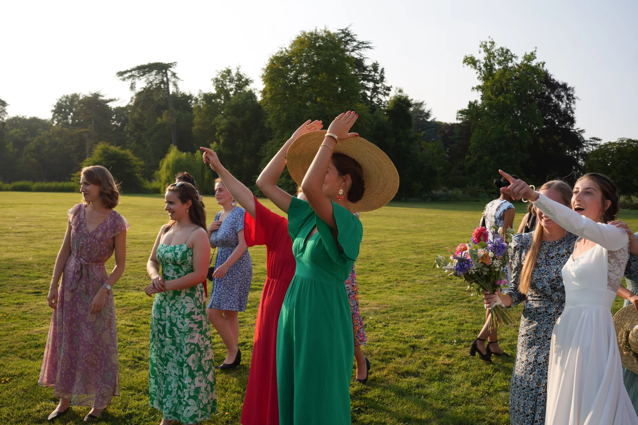 Un groupe de femmes en robes estivales participent à une cérémonie en plein air, certaines tenant des bouquets de fleurs, avec des arbres en arrière-plan.