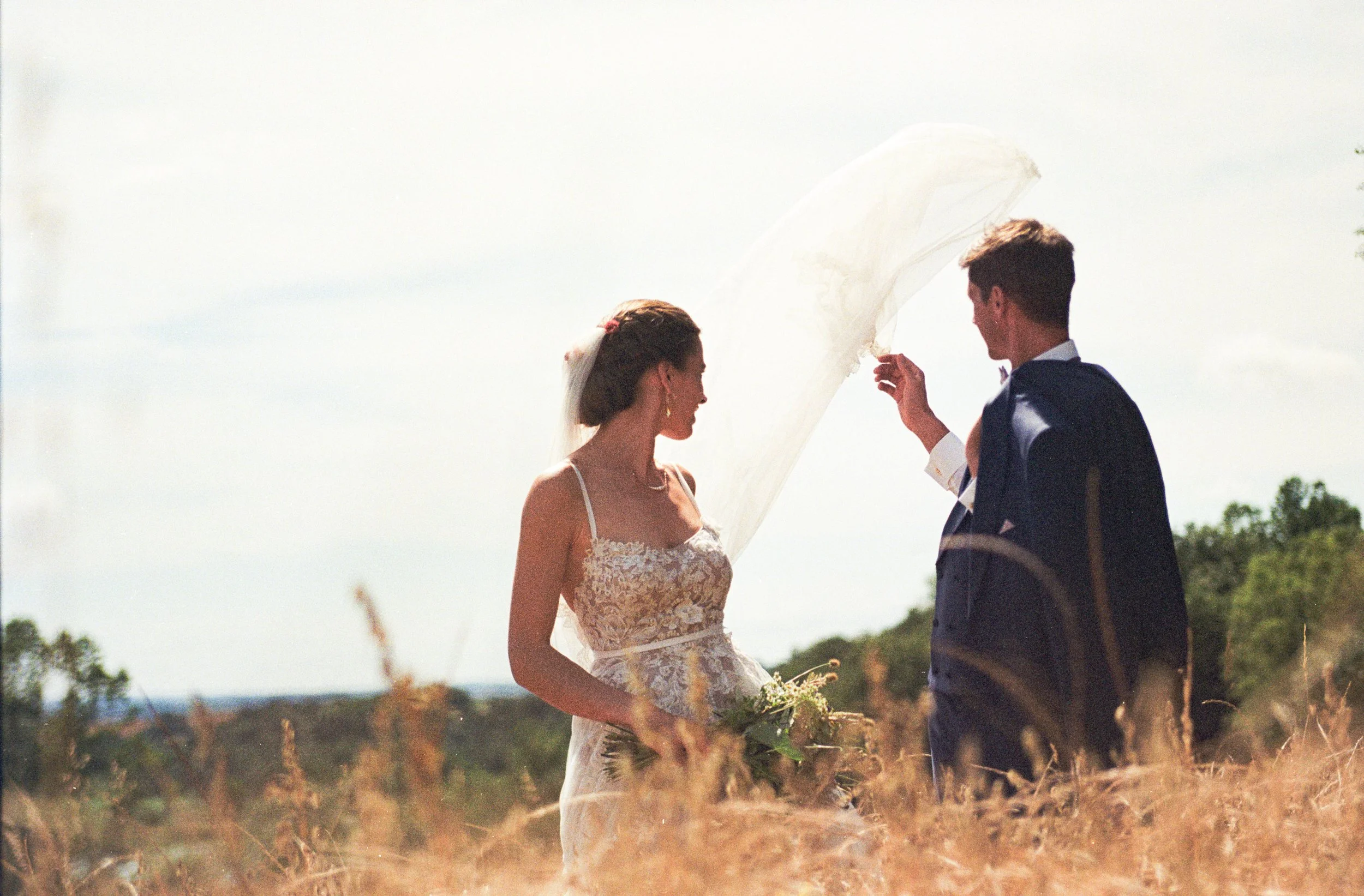 Un couple de mariés se tenant la main dans un champ en plein air, la femme en robe de mariage avec un voile, l'homme en costume, sous un parapluie blanc, en arrière-plan un ciel clair et un paysage naturel.
