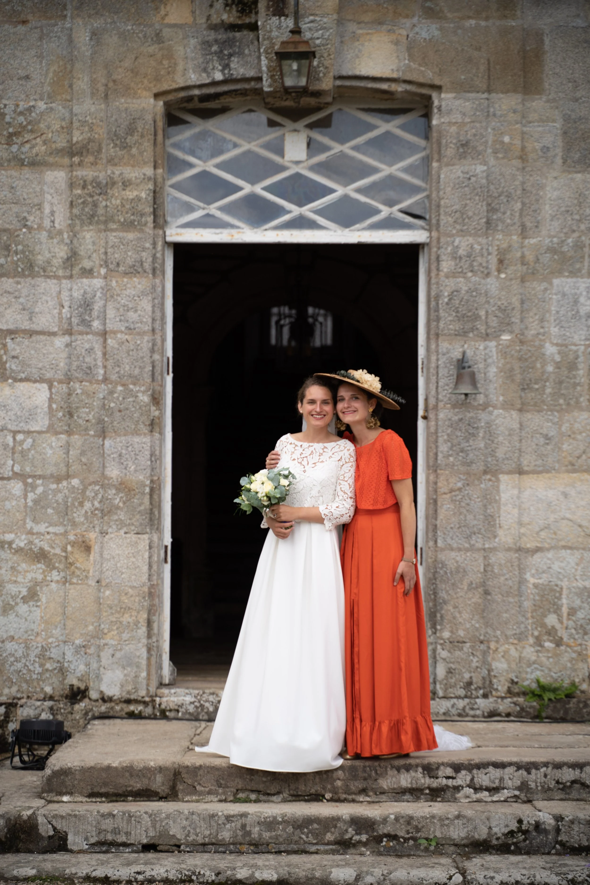 Deux femmes souriantes se tiennent sous une arche en pierre, l'une en robe blanche de mariée tenant un bouquet de fleurs, l'autre en robe orange avec un grand chapeau, devant un bâtiment ancien.