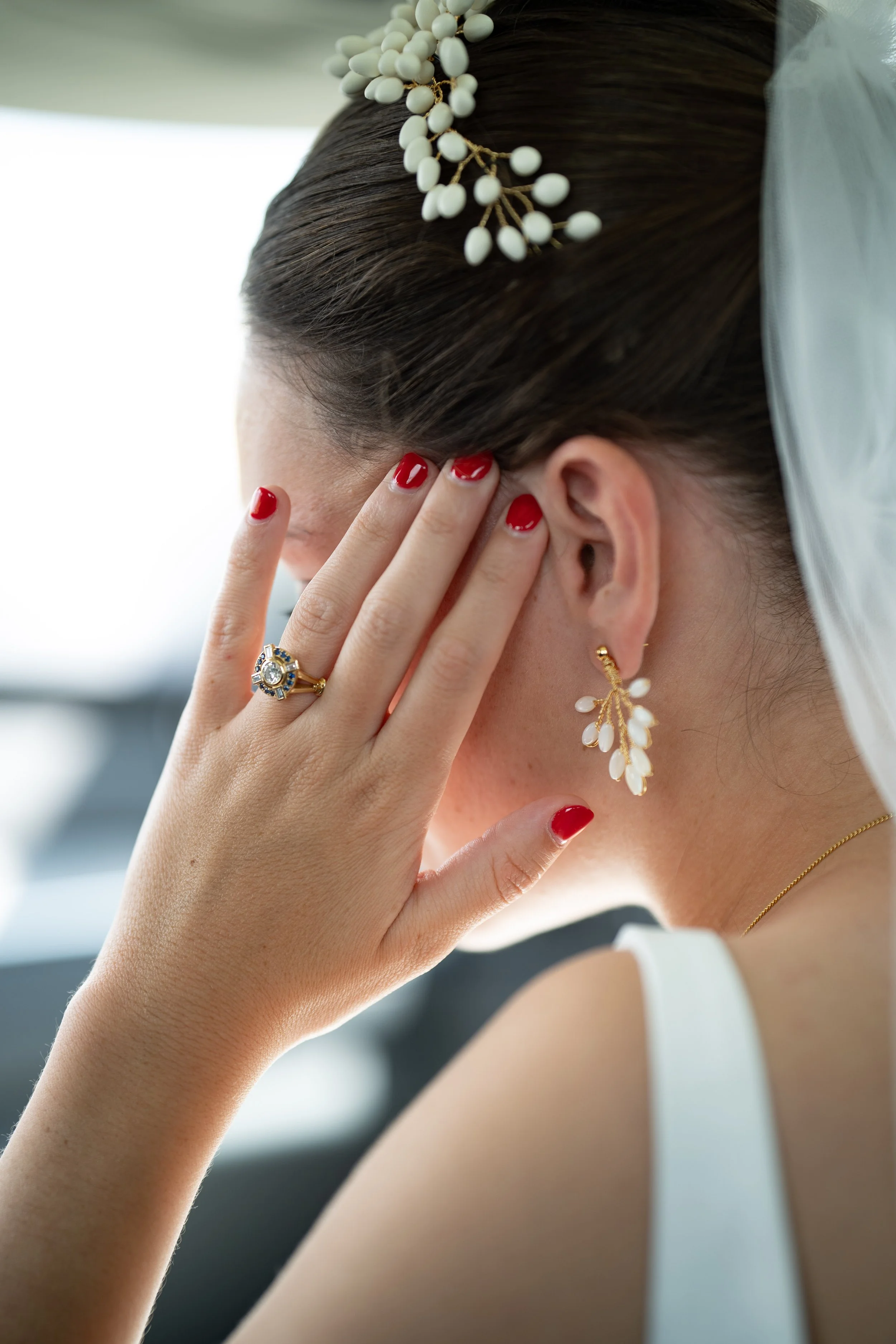 Une femme avec des cheveux foncés, portant des boucles d'oreilles élégantes et un bijou sur la tête, se tenant la main sur le visage, avec des ongles rouge vif et une bague ornementale. Elle porte une robe blanche et une fine chaîne dorée.