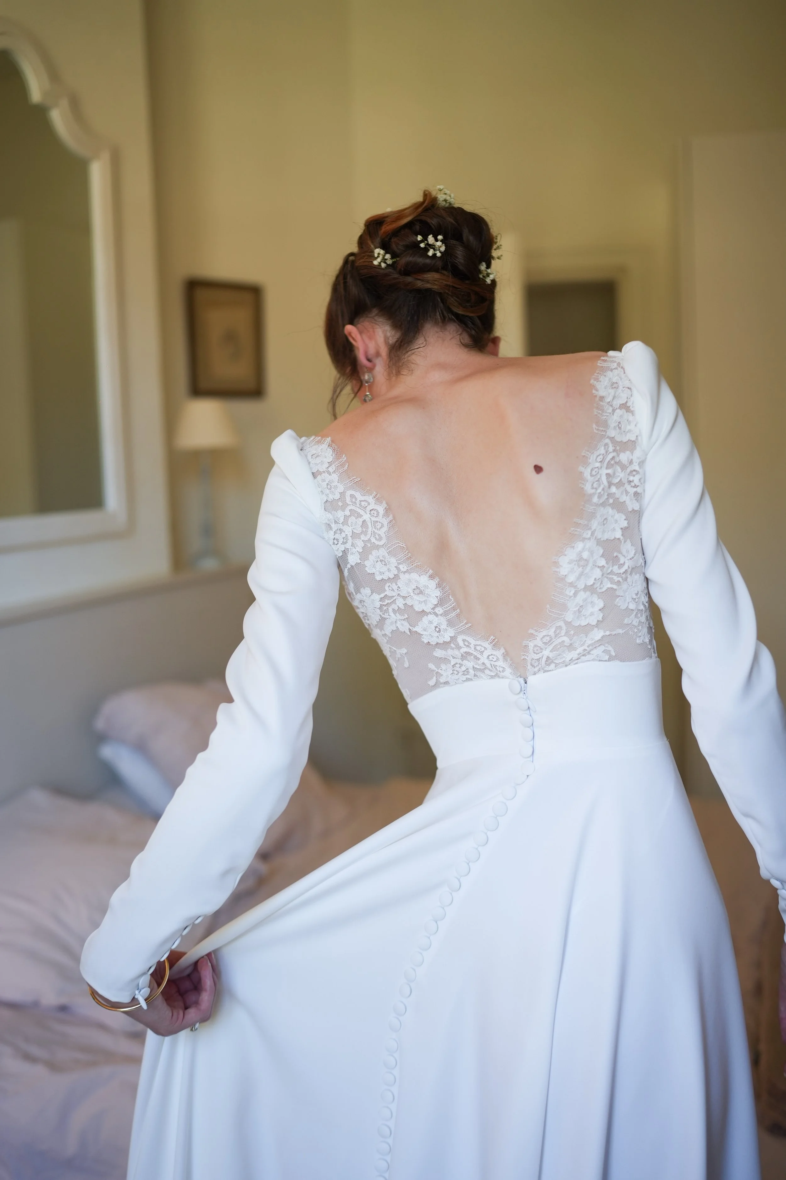 Une femme en robe de mariée ajustant la couture de son dos avec des boutons. Cheveux coiffés avec des fleurs, boucles d'oreilles pendantes dans une pièce chaleureuse.