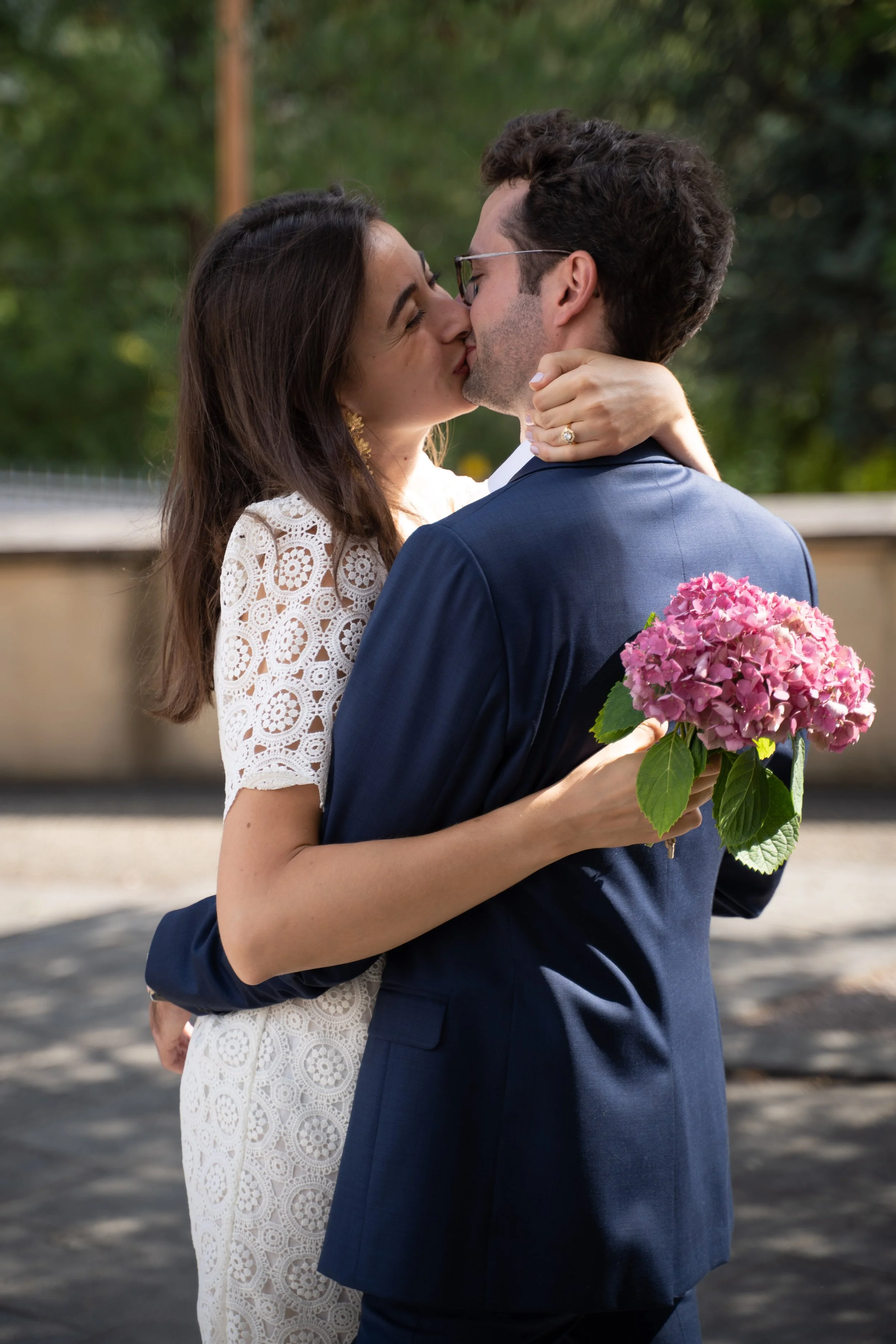 Un couple s'embrasse lors de leur mariage, la femme porte une robe blanche en dentelle et tient un bouquet de hortensias roses.
