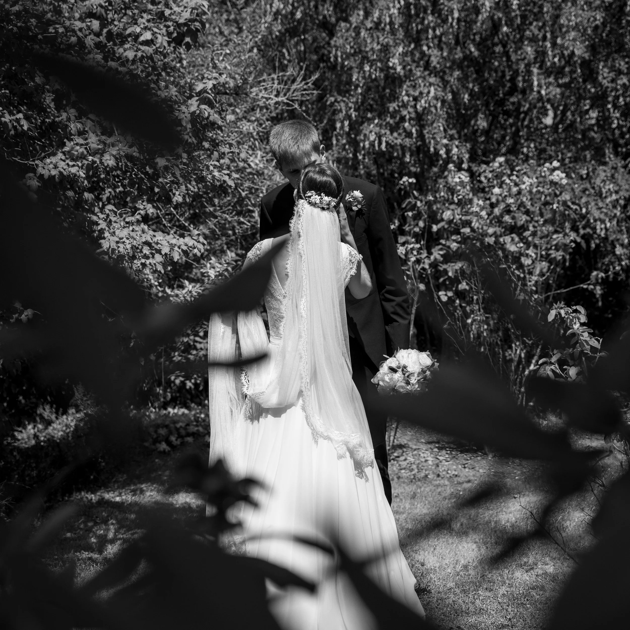 Un couple de mariés marchant dans la nature, vue de dos, entourés de végétation dense, en noir et blanc.