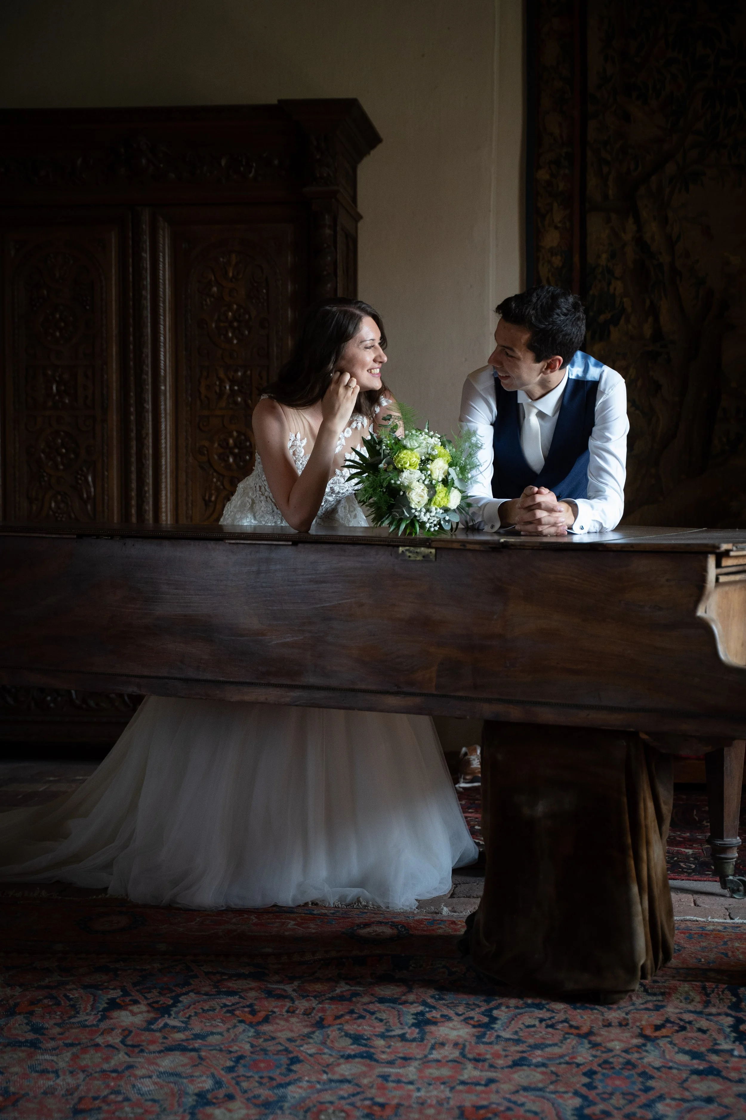 Un couple marié souriant et regardant l'un l'autre, assis derrière un vieux piano en bois recouvert d'une robe blanche, avec un bouquet de fleurs blanches et vertes sur le piano, dans une pièce avec un décor vintage et un tapis persan.
