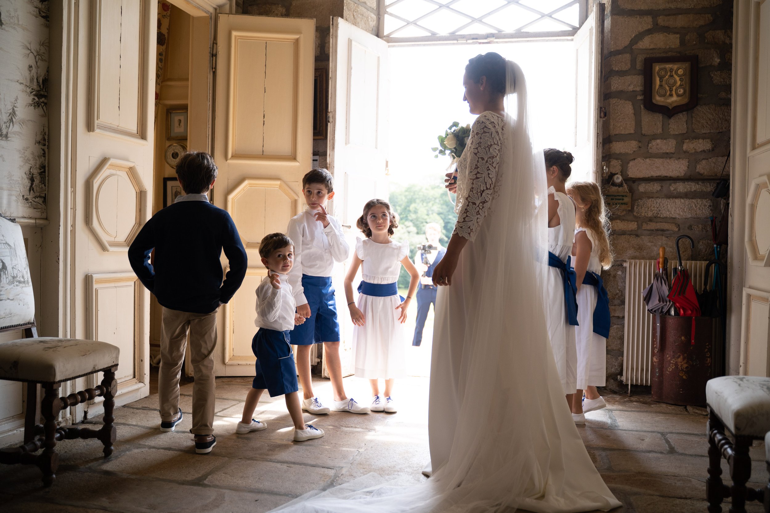 Une mariée souriante en robe blanche tenant un bouquet de fleurs, entourée d'enfants en tenue blanche et bleue, dans une pièce lumineuse avec une porte ouverte donnant sur l'extérieur.