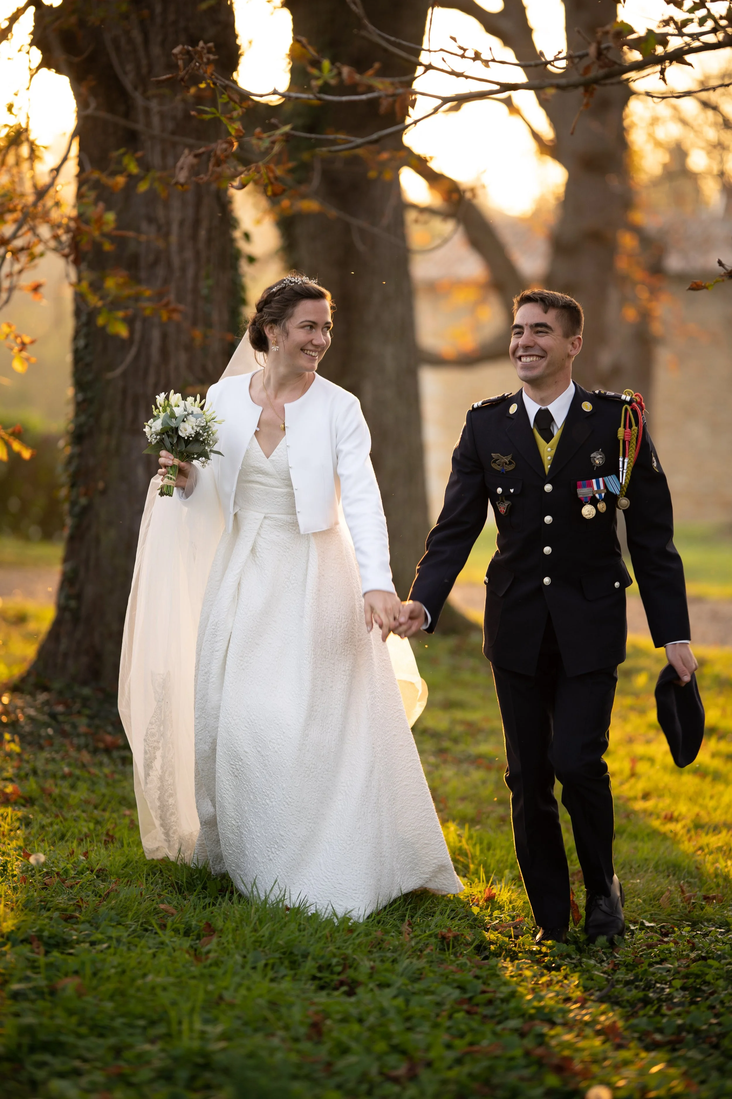 Un couple de mariés, une femme en robe de mariage blanche et un homme en uniforme militaire, marchent ensemble dans un parc lors du coucher du soleil, tenant la main et sourire