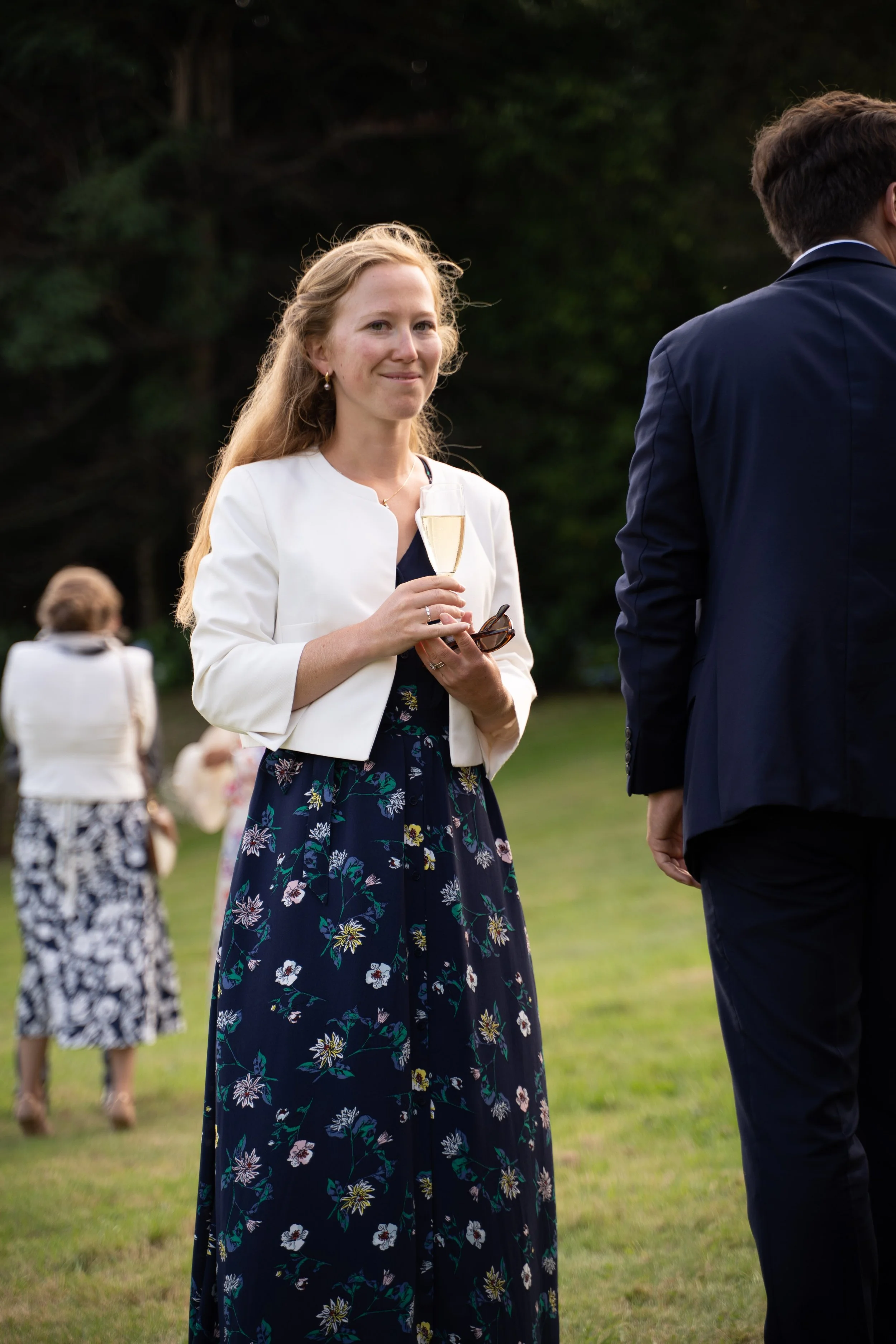 Une femme souriante tenant un verre de champagne lors d'une réception en plein air, portant une robe longue à motifs floraux et une veste blanche, avec un environnement verdoyant en arrière-plan.