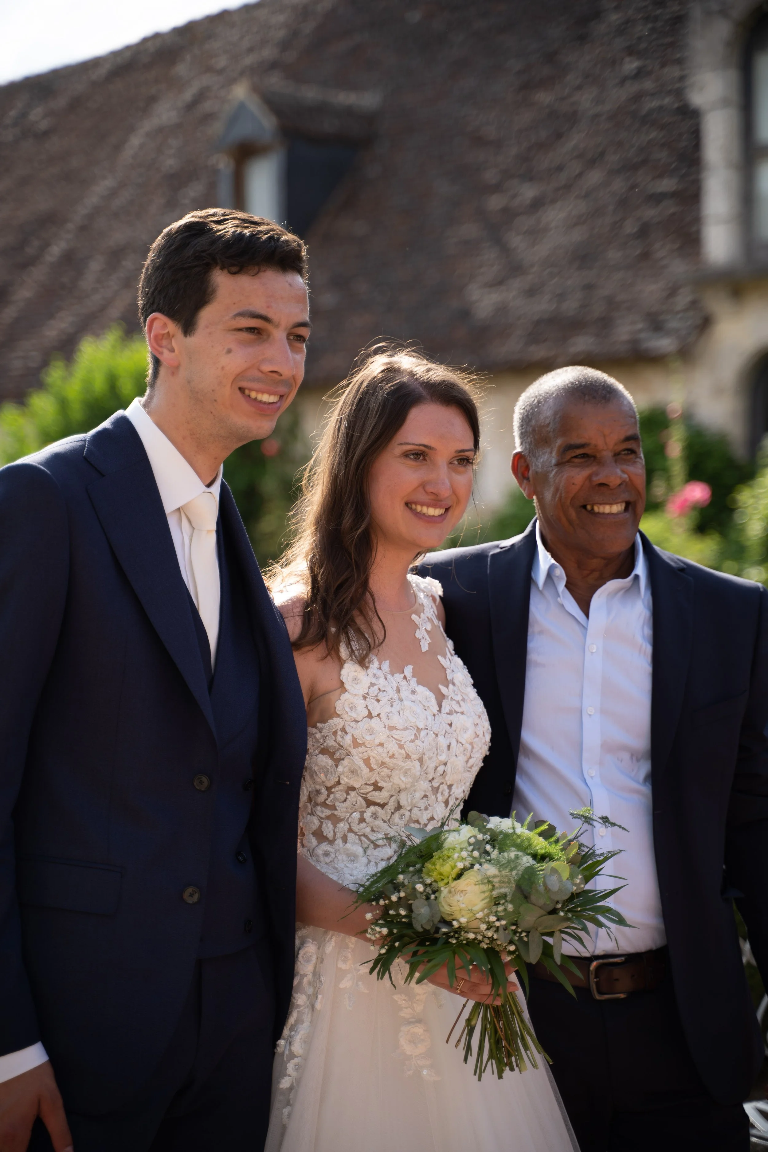 Trois personnes lors d'un mariage, un homme en costume noir, une femme en robe de mariée blanche et un homme en costume sombre, souriant, avec un gâteau ou bouquet à la main, devant une maison en pierre et un jardin.