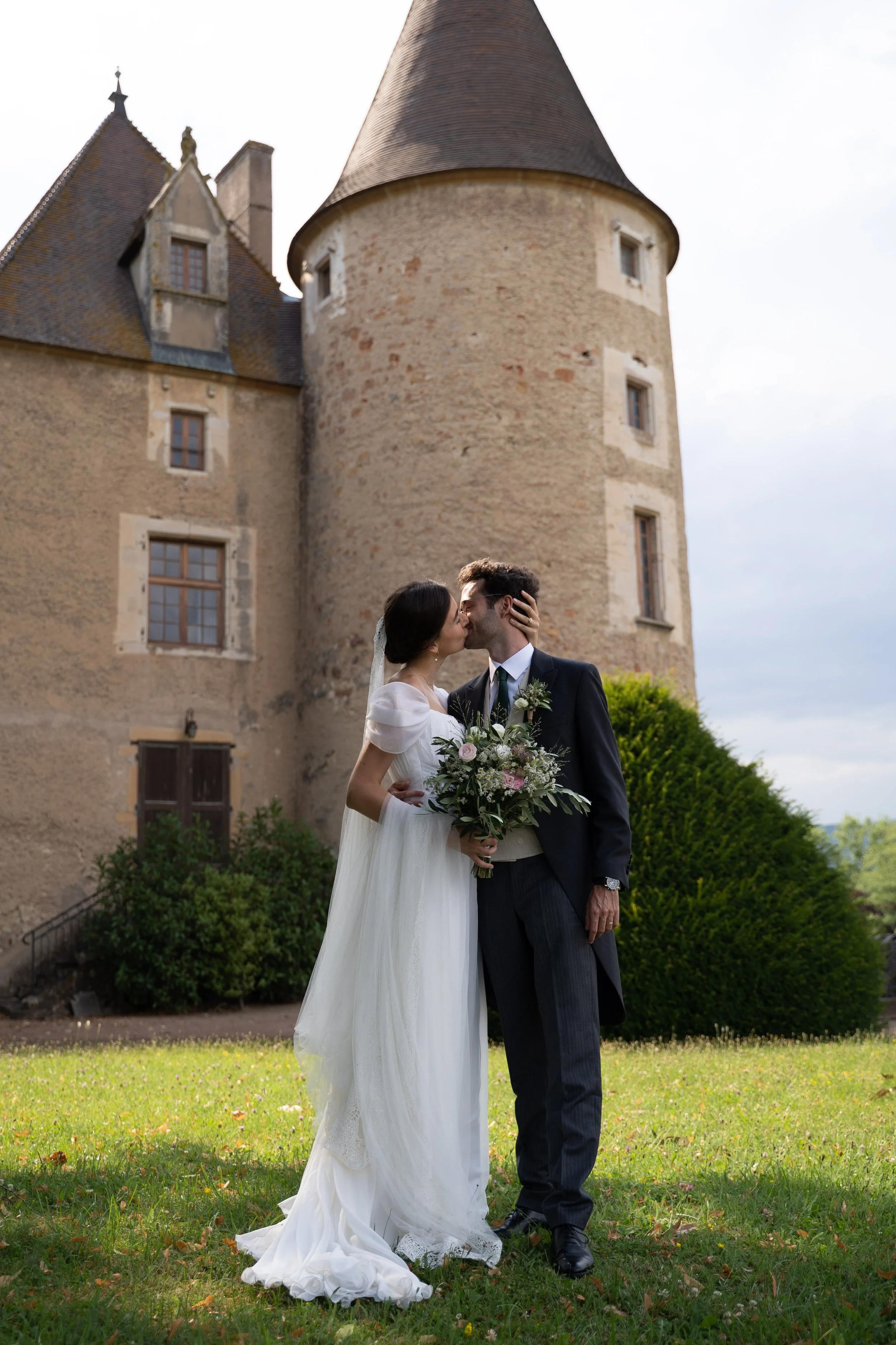 Un couple de mariés s'embrassent devant un château avec des murs en pierre et des fenêtres en bois, dans un jardin vert.