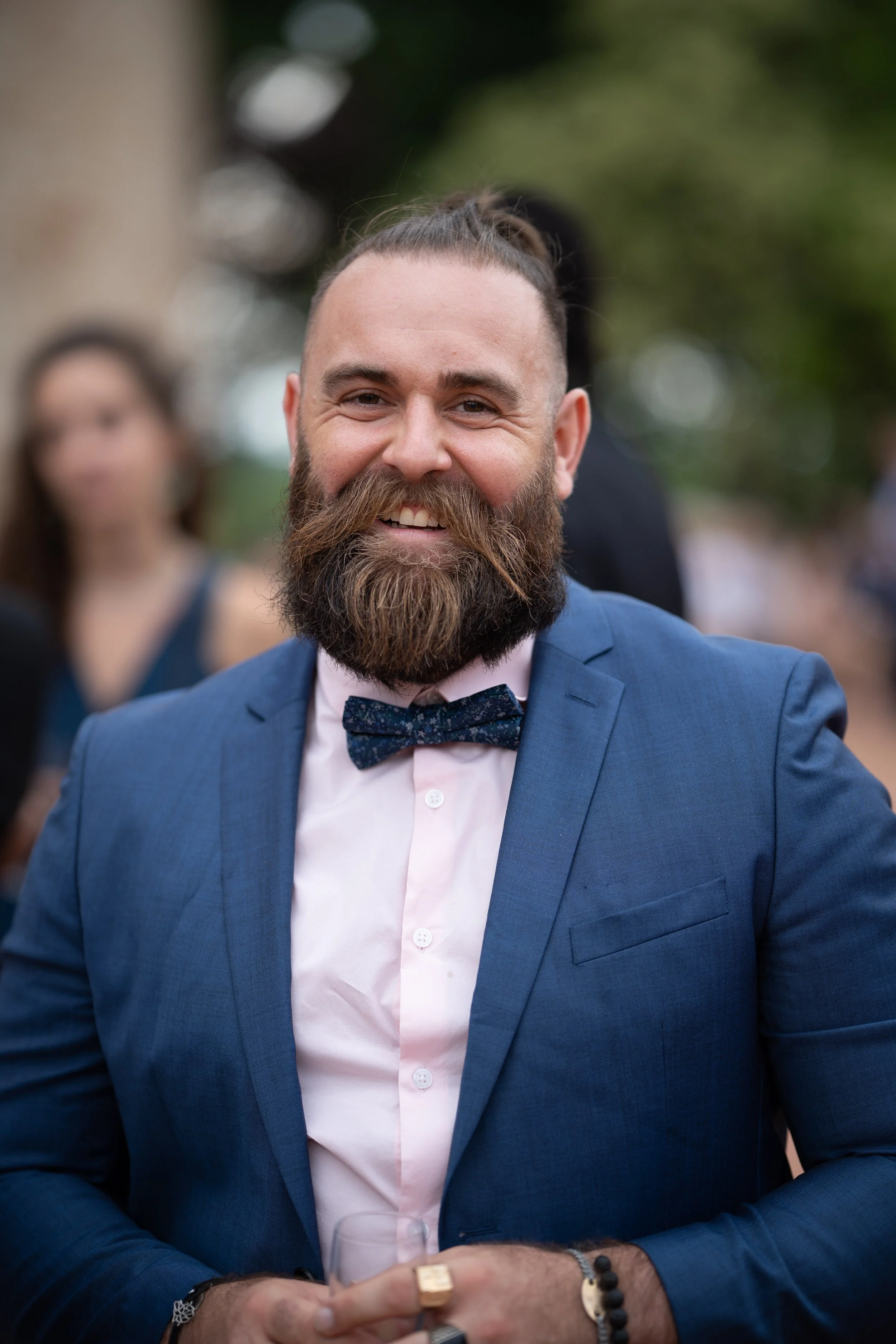 Un homme souriant portant un costume bleu, une chemise blanche et un nœud papillon noir à motifs, lors d'un événement en plein air. Il a une barbe épaisse et porte plusieurs bracelets et une bague.