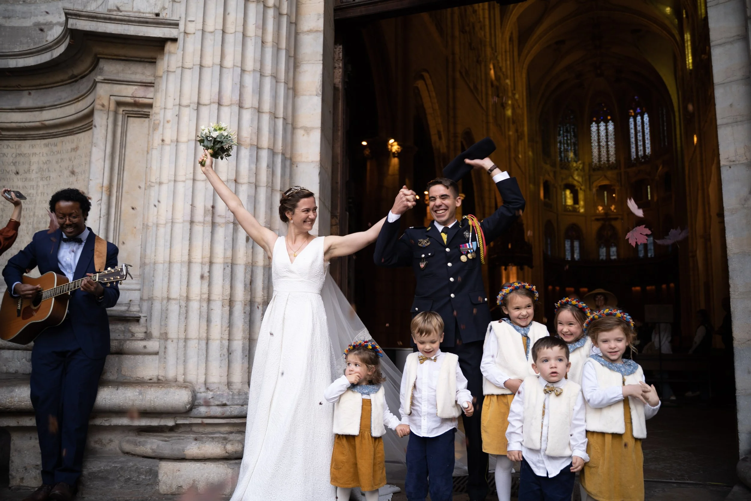 Un groupe de personnes, principalement des enfants en vêtements traditionnels, dans une église ou une cathédrale, avec une femme en robe de mariage et un homme en uniforme militaire, célébrant un mariage.