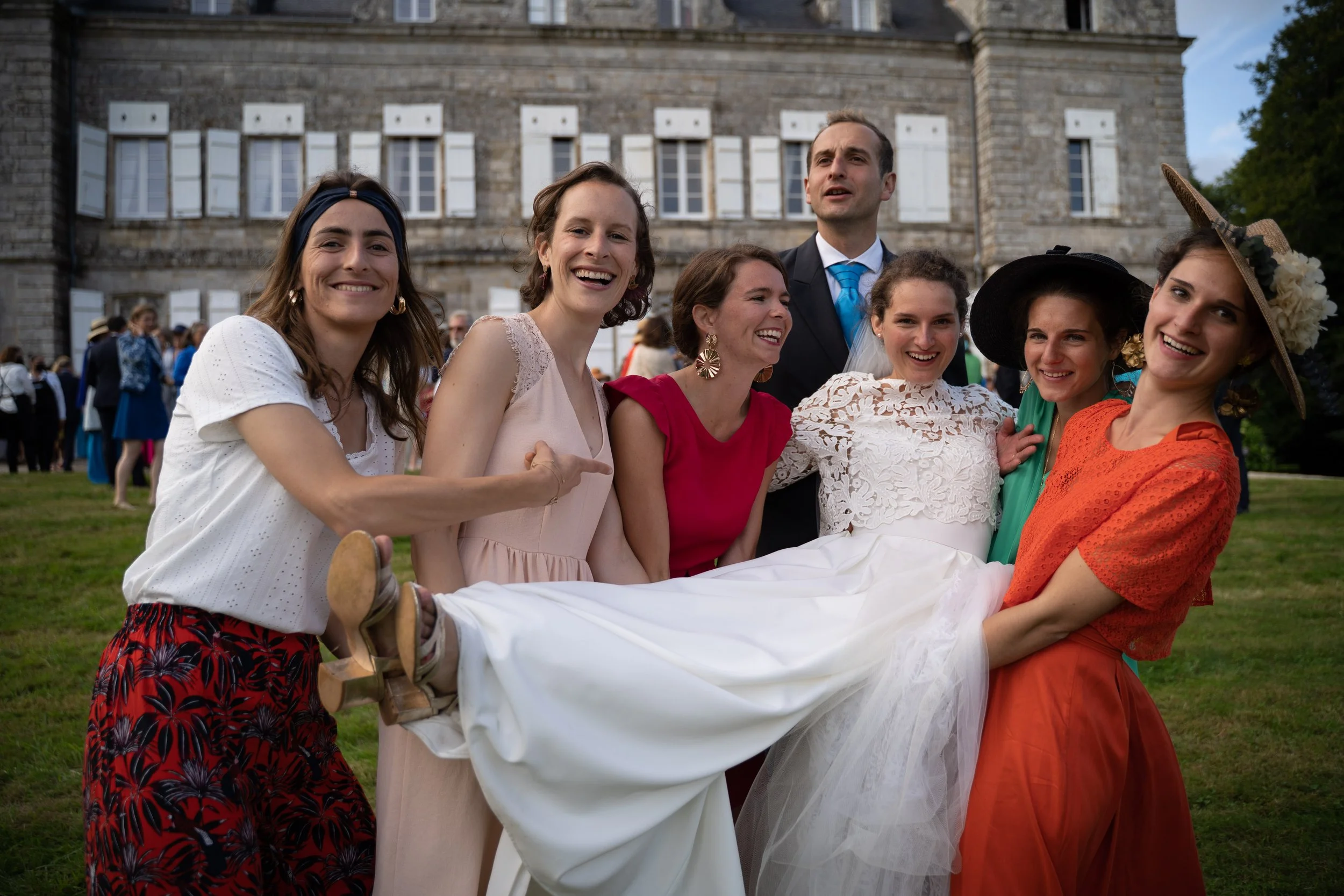 Groupe de personnes souriantes portant une mariée allongée lors d'une célébration en extérieur devant un grand bâtiment en pierre.