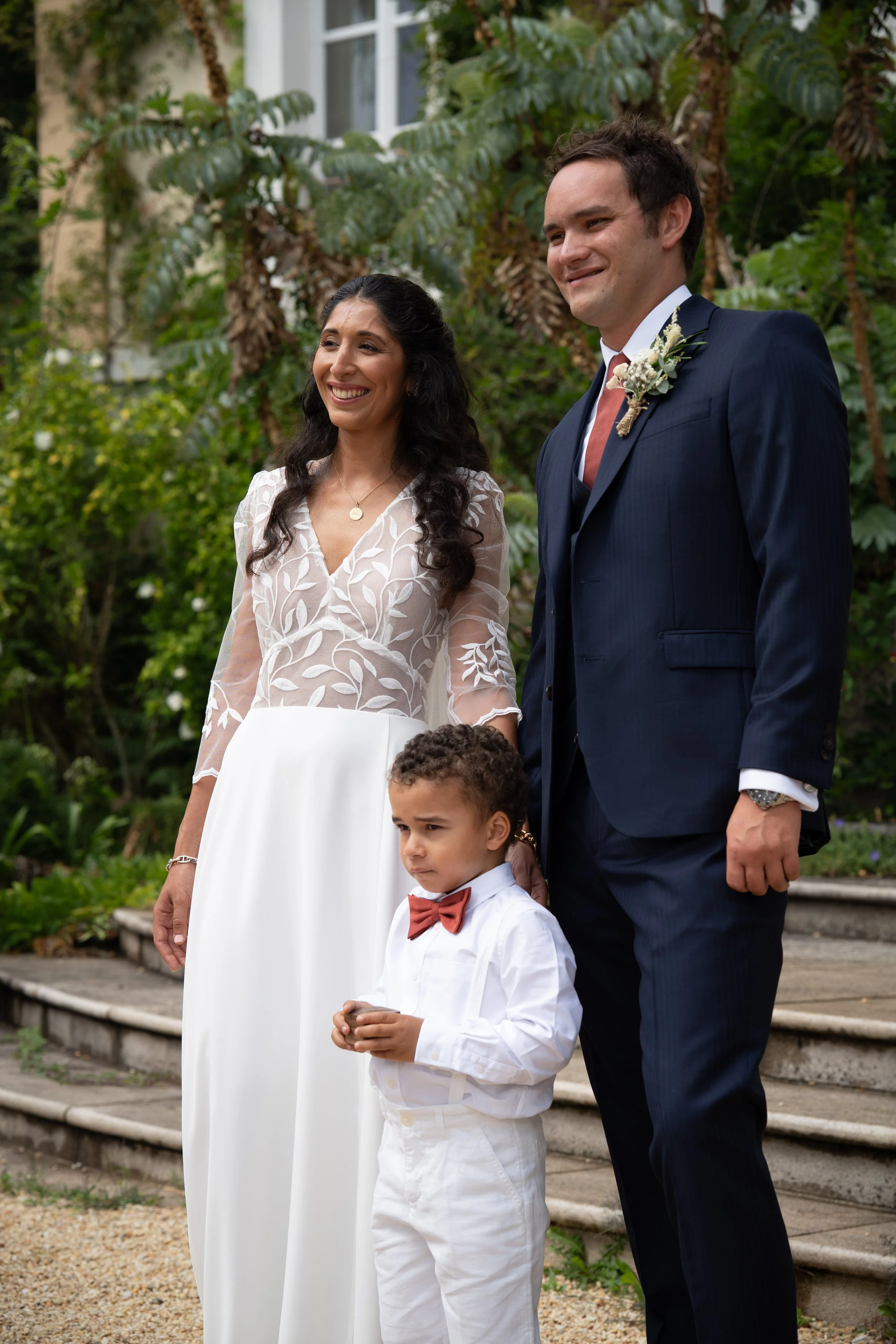 Un couple vêtu pour un mariage, avec un enfant, posant à l'extérieur dans un jardin avec des buissons et des arbres.