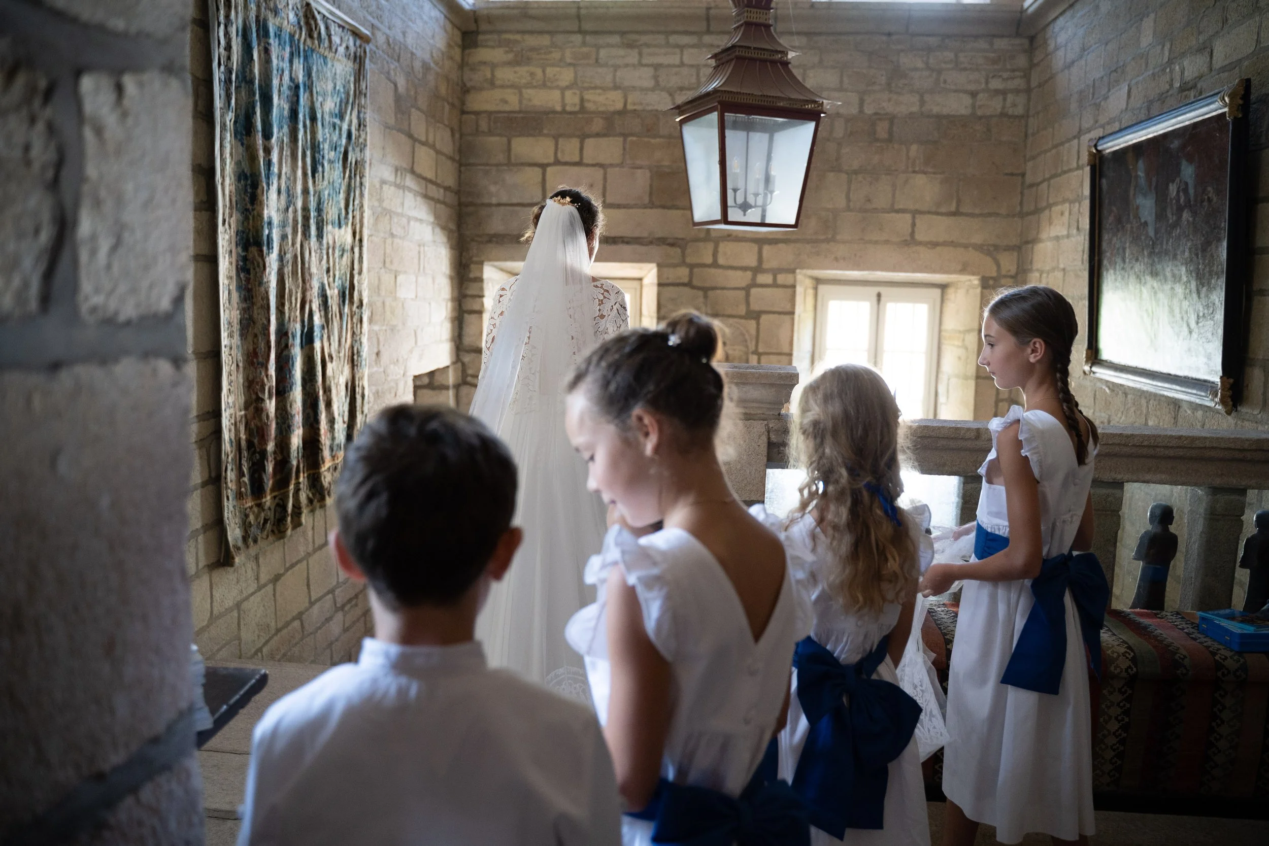 Une jeune fille en robe blanche avec un voile participe à une cérémonie religieuse dans une salle en pierre, entourée d'autres enfants.
