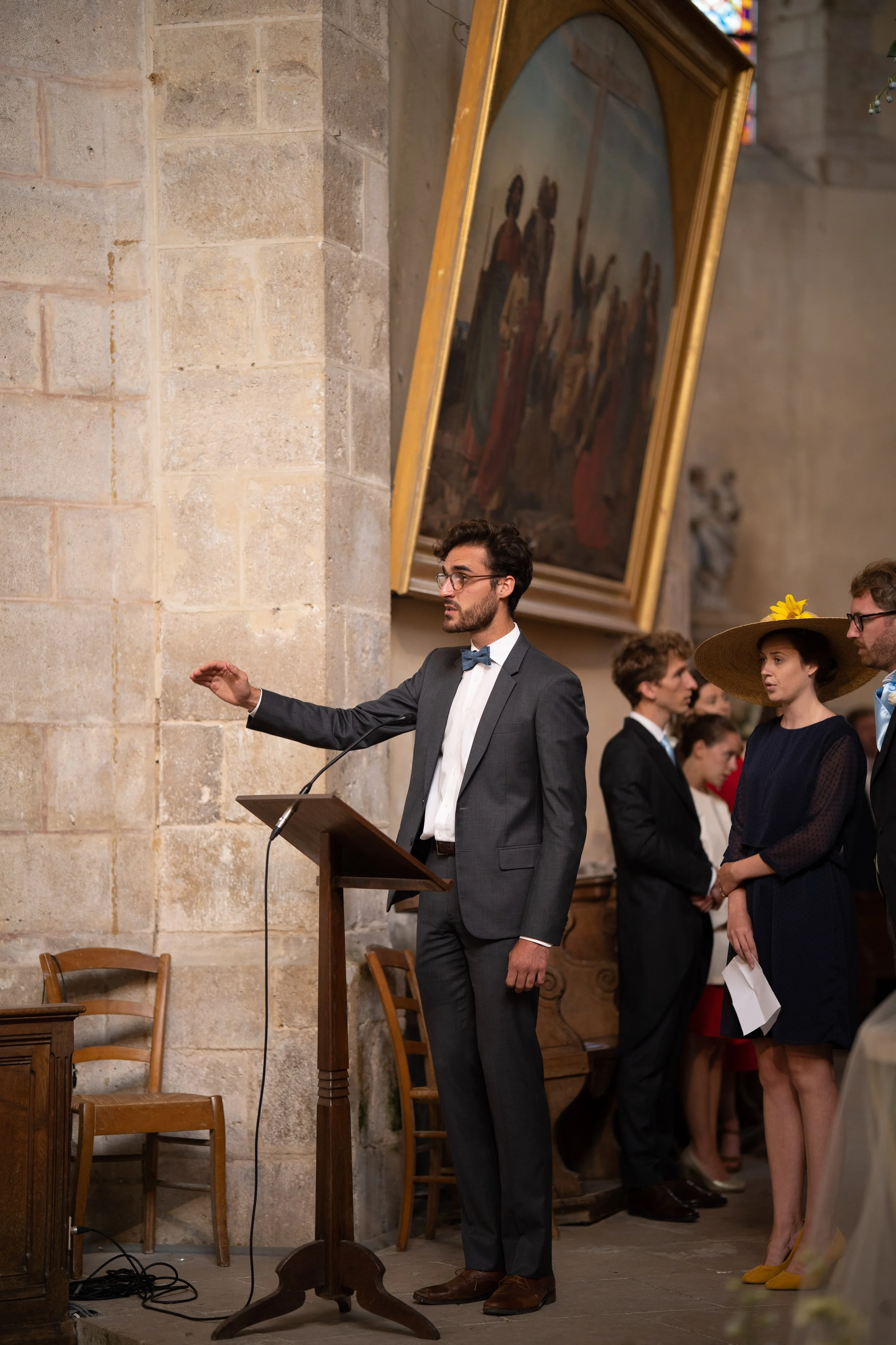Un homme en costume gris et cravate noire parle à un micro devant une foule dans une église en pierre avec un tableau religieux en arrière-plan. D'autres personnes en vêtements formels attendent ou écoutent dans la même pièce.