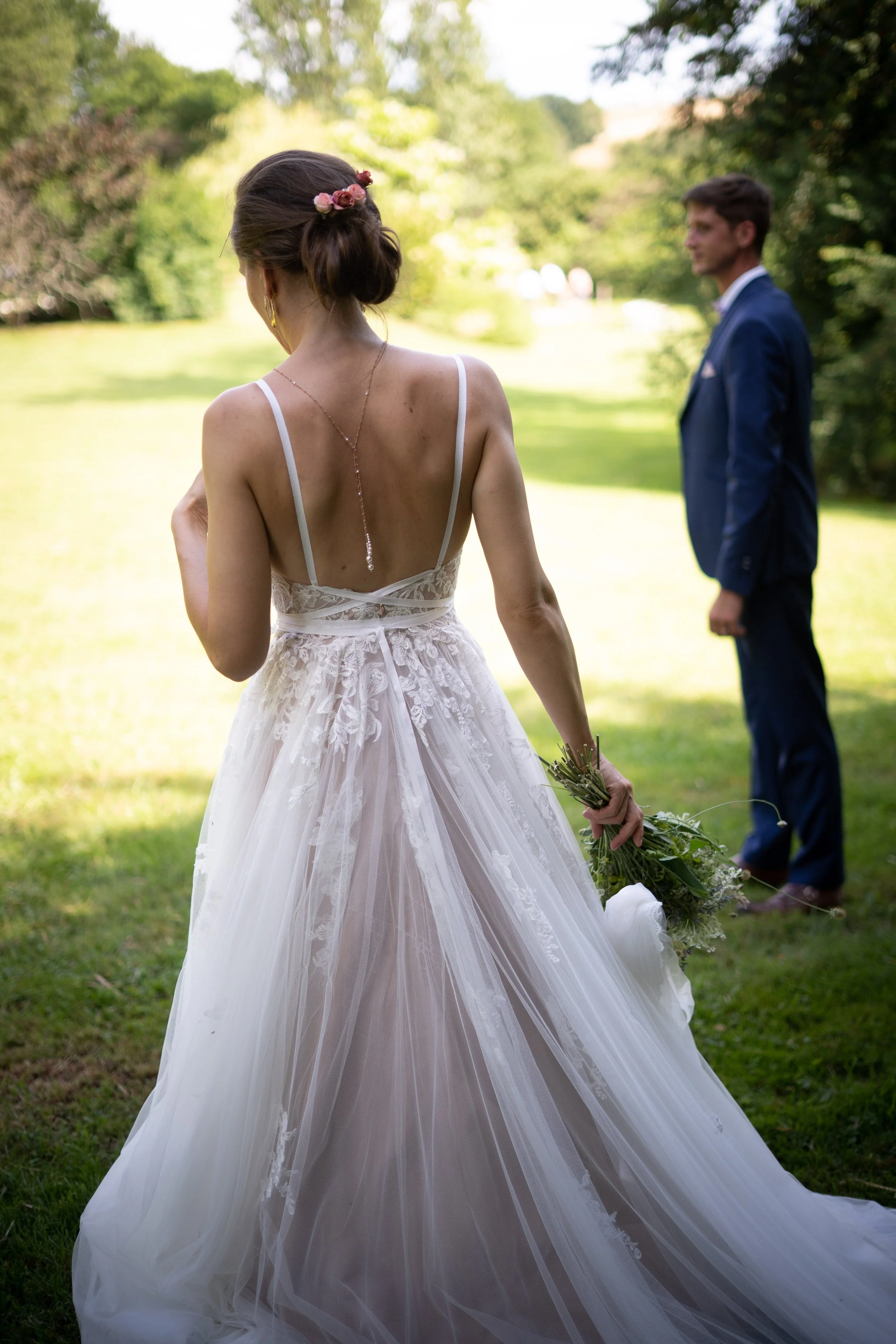 Une mariée en robe de mariage blanche avec un bouquet, vue de dos, dans un parc en plein air, avec un homme en costume en arrière-plan.