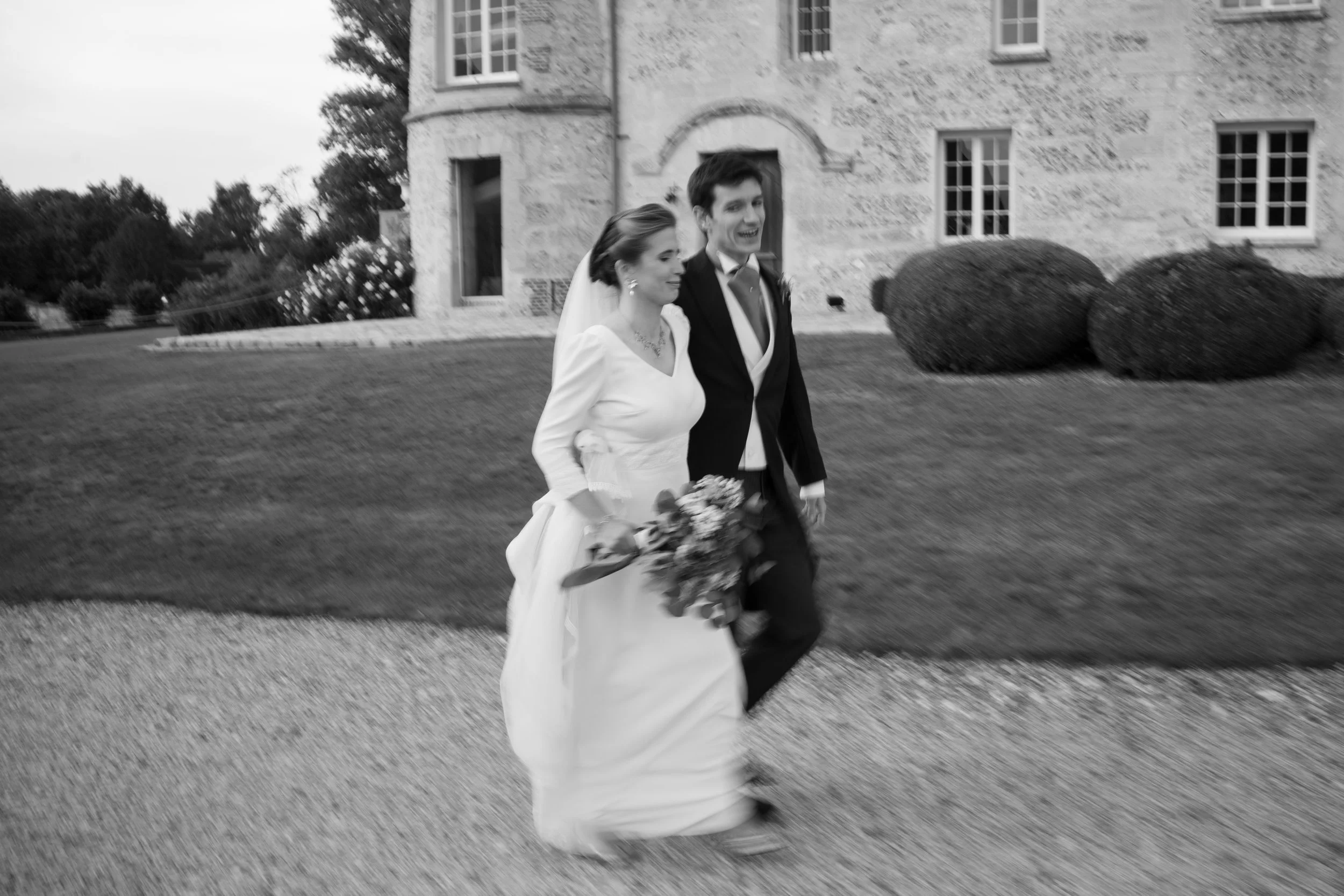 Un couple de mariés marche devant une grande maison en pierre, sourire et élégance, la mariée porte une robe blanche et le marié un costume noir.