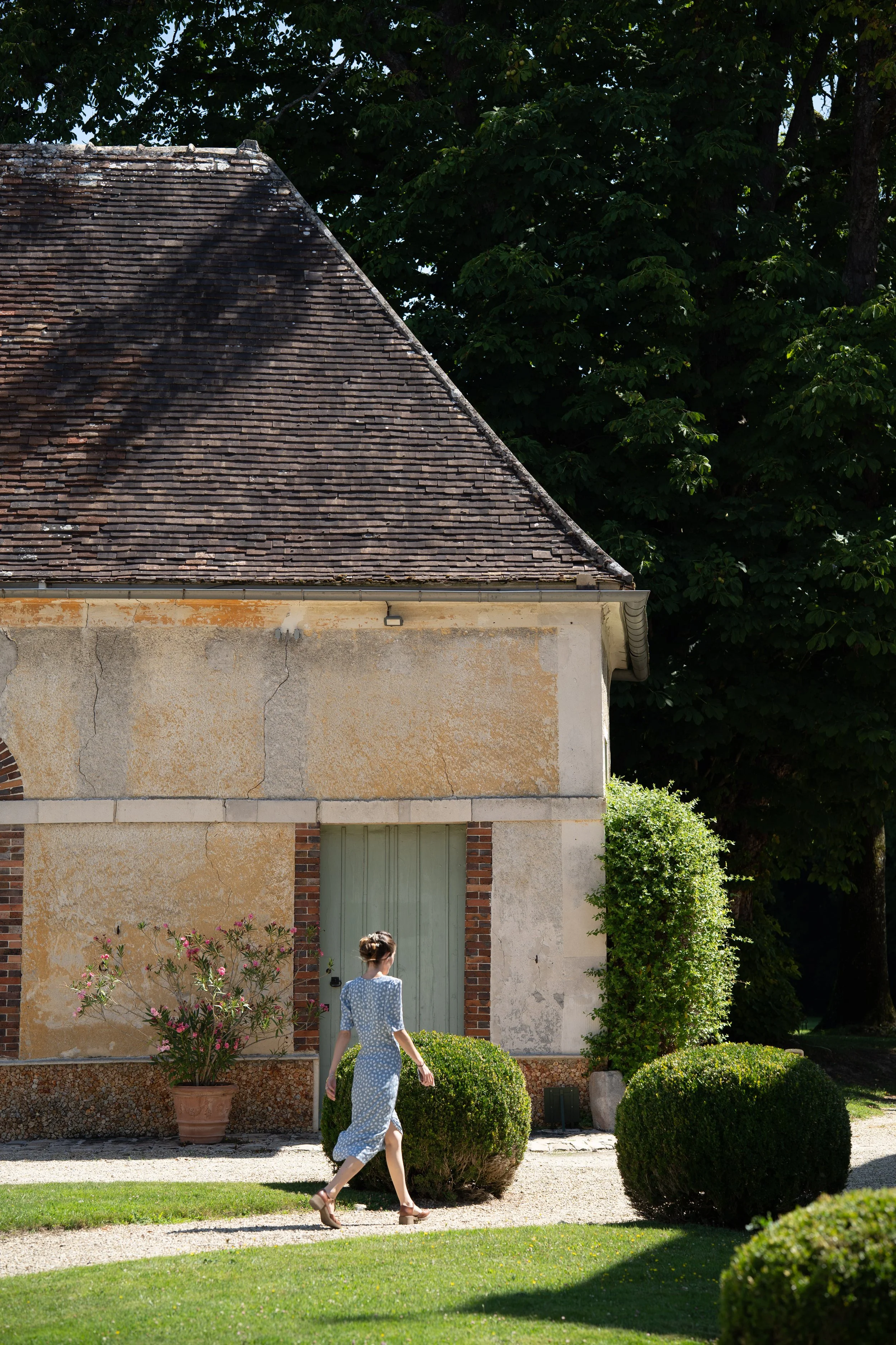 Une femme en robe bleue à pois blancs marche devant une maison ancienne avec un toit en tuiles, entourée de buissons et d'arbres verts.