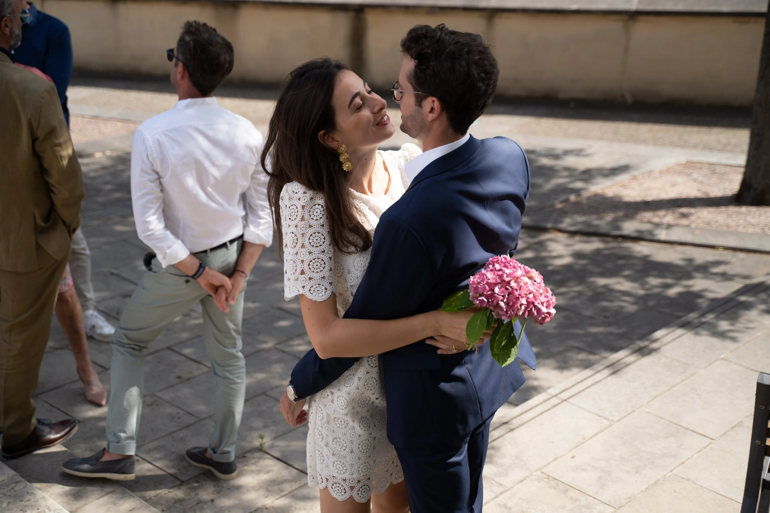 Un couple en habits élégants se tenant dans une embrassade, la femme tenant un bouquet de fleurs roses lors d'un événement en plein air.