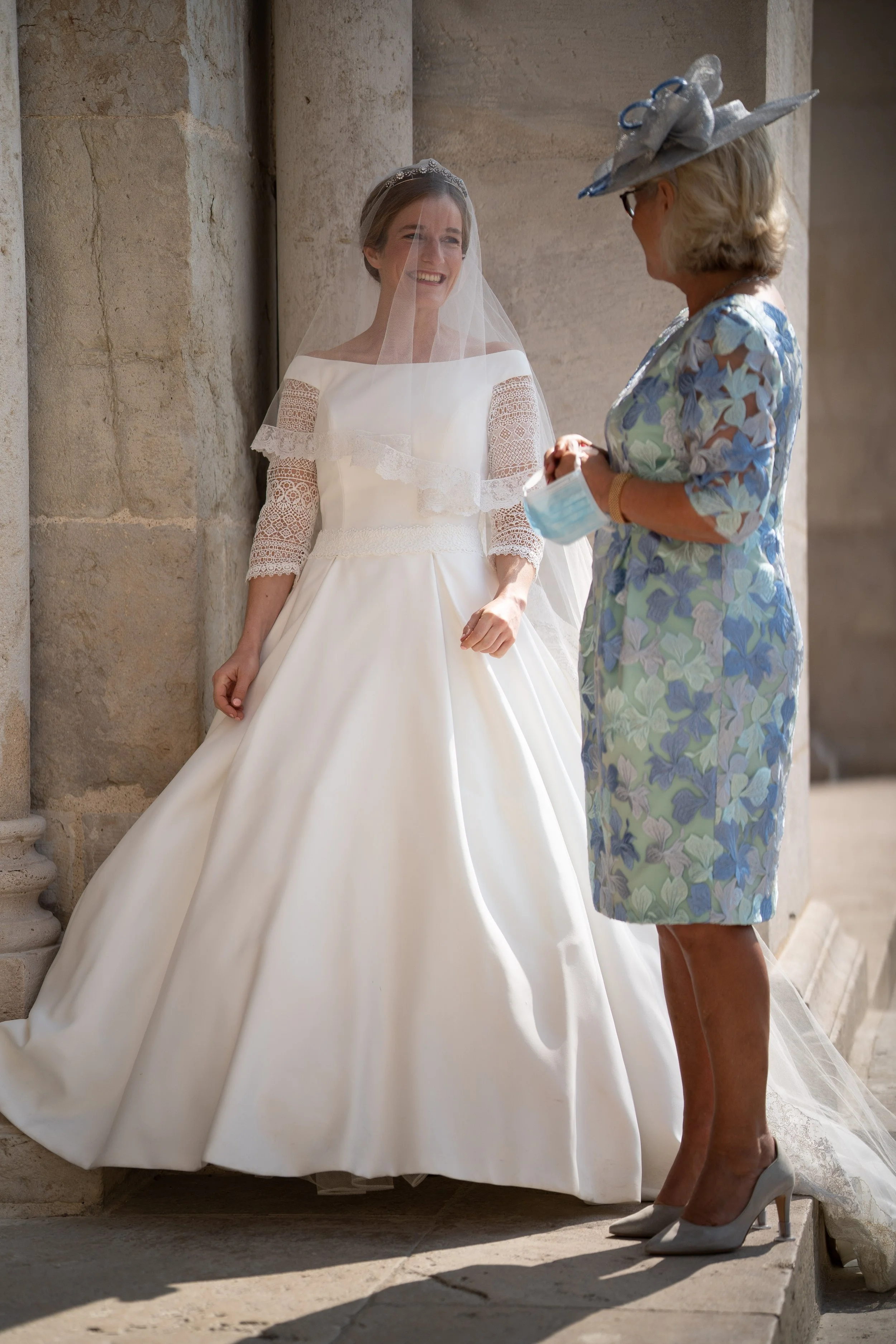 Photo d'une mariée avec une robe blanche longue et un voile transparent, parlant avec une femme âgée portant un chapeau décoré et une robe à motifs floraux de couleurs pastel, toutes deux debout devant un mur en pierre.