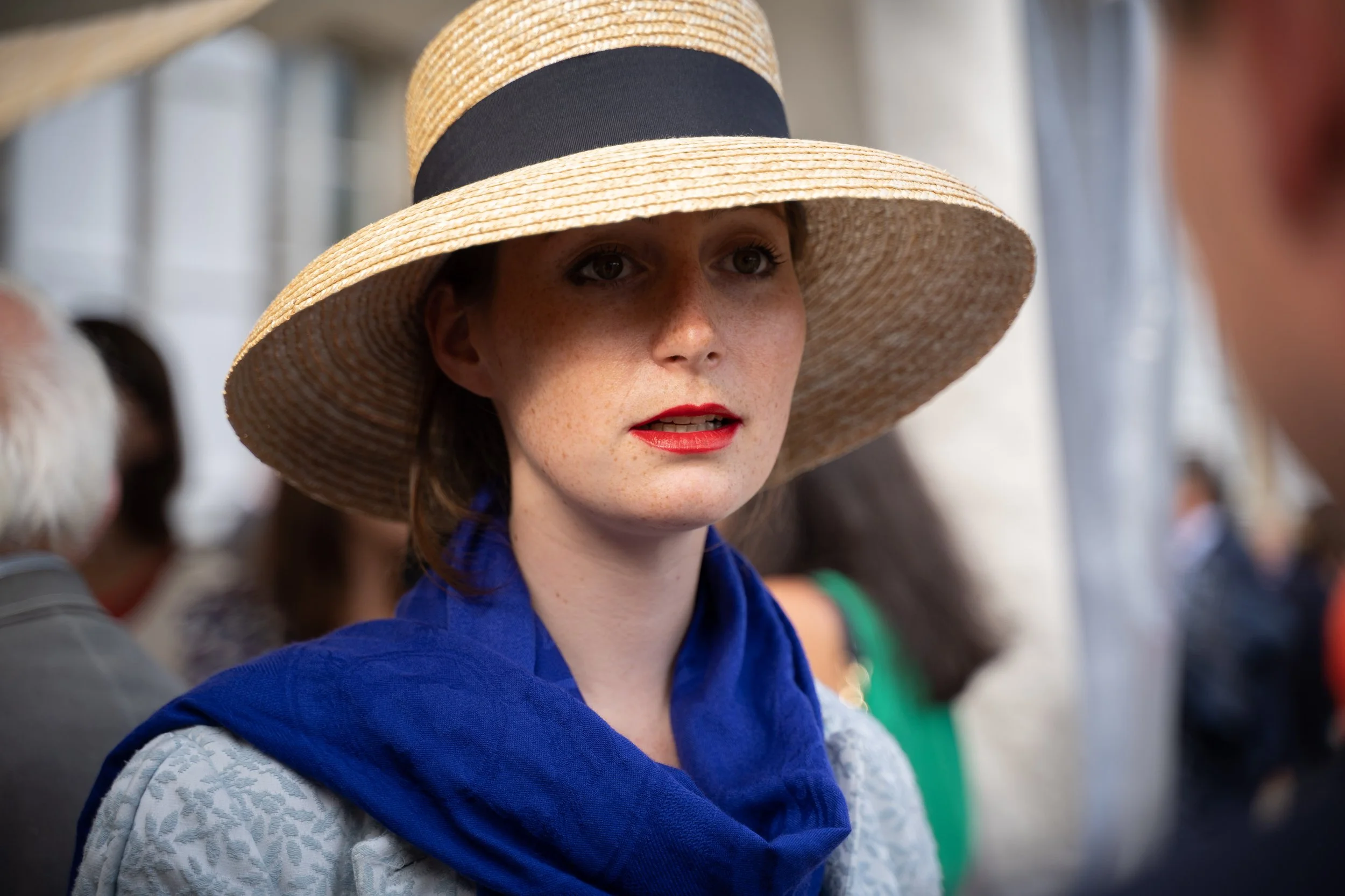 Femme portant un chapeau de paille avec un ruban noir, vêtue d'une veste bleue, en discussion avec quelqu'un dans un environnement extérieur.