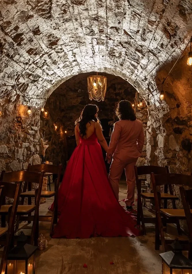 A woman in a long red evening gown and a man in a beige suit holding hands, standing in a rustic stone-walled room with warm lighting and wooden chairs.