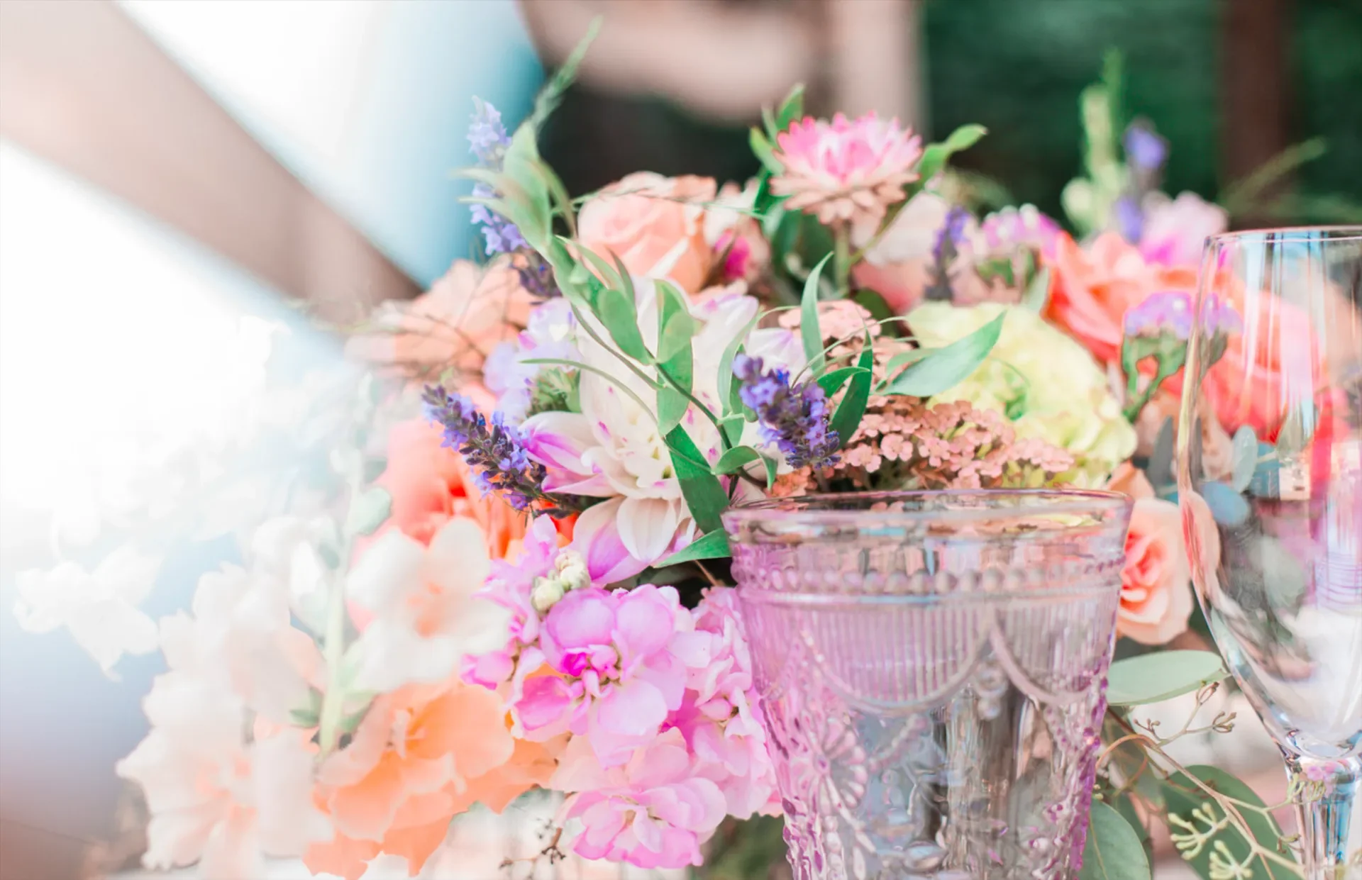 Close-up of a floral centerpiece with pink, white, and purple flowers on a table, with two glassware cups in foreground.