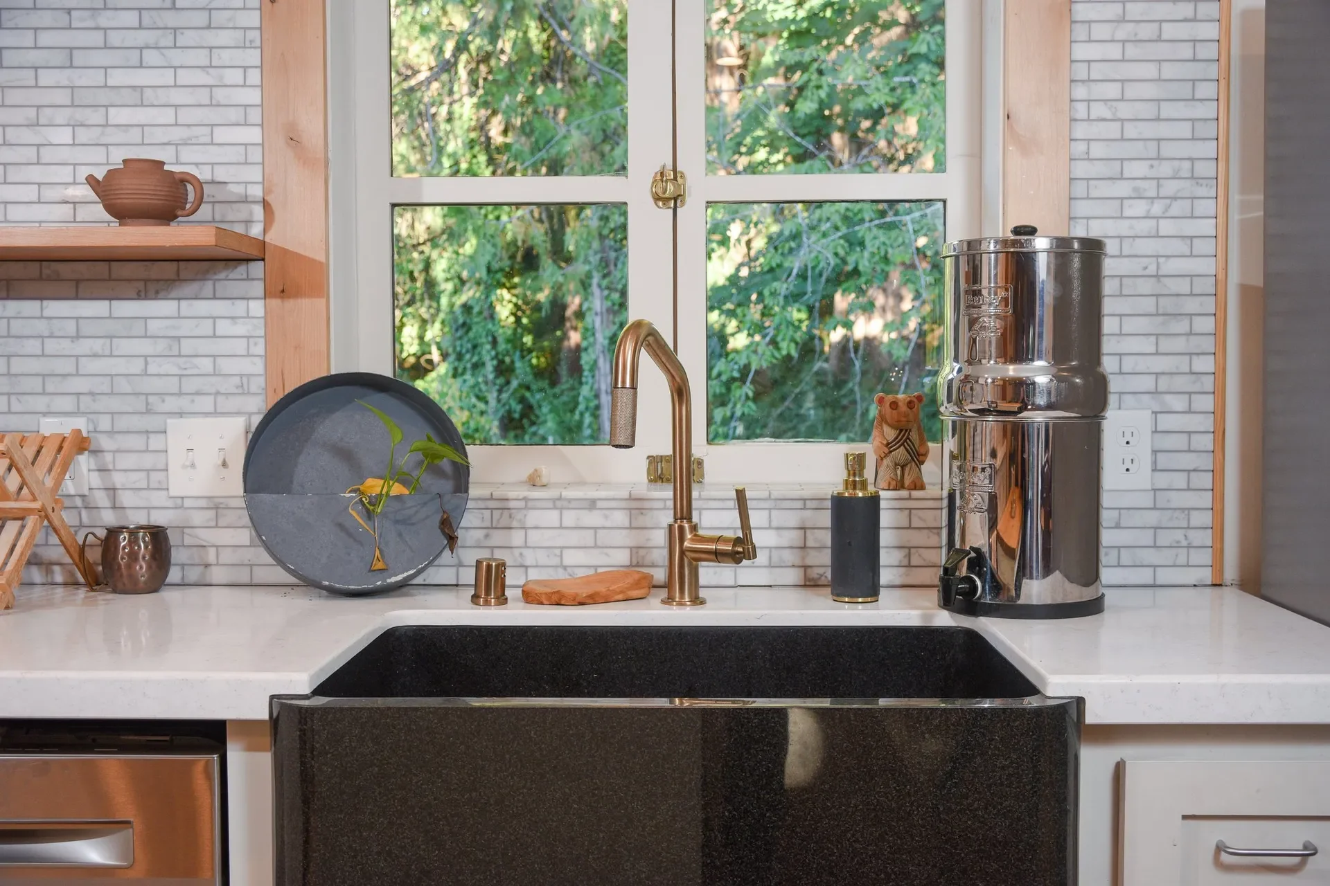 Kitchen sink with gold faucet, soap dispenser, decorative bear figurine, water filter, and window showing green trees outside.