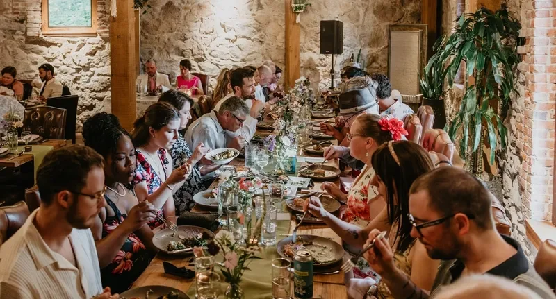 People dining together at a long banquet table in a rustic restaurant with stone walls and large window, enjoying a meal and conversation.